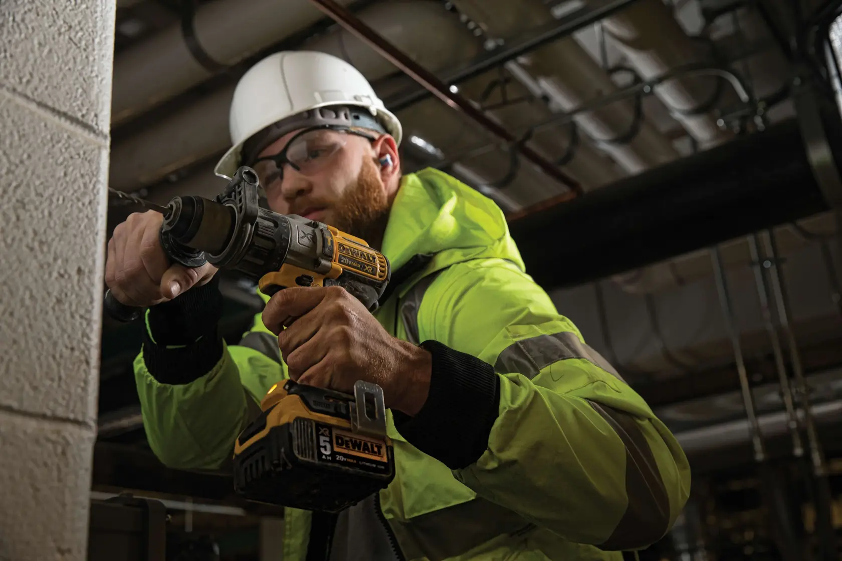 A person wearing a high-visibility jacket and hard hat is using a DEWALT cordless power drill (model DCD996P2) to drill into a concrete or masonry wall in an industrial or construction setting. The product and grip on the tool are clearly visible.