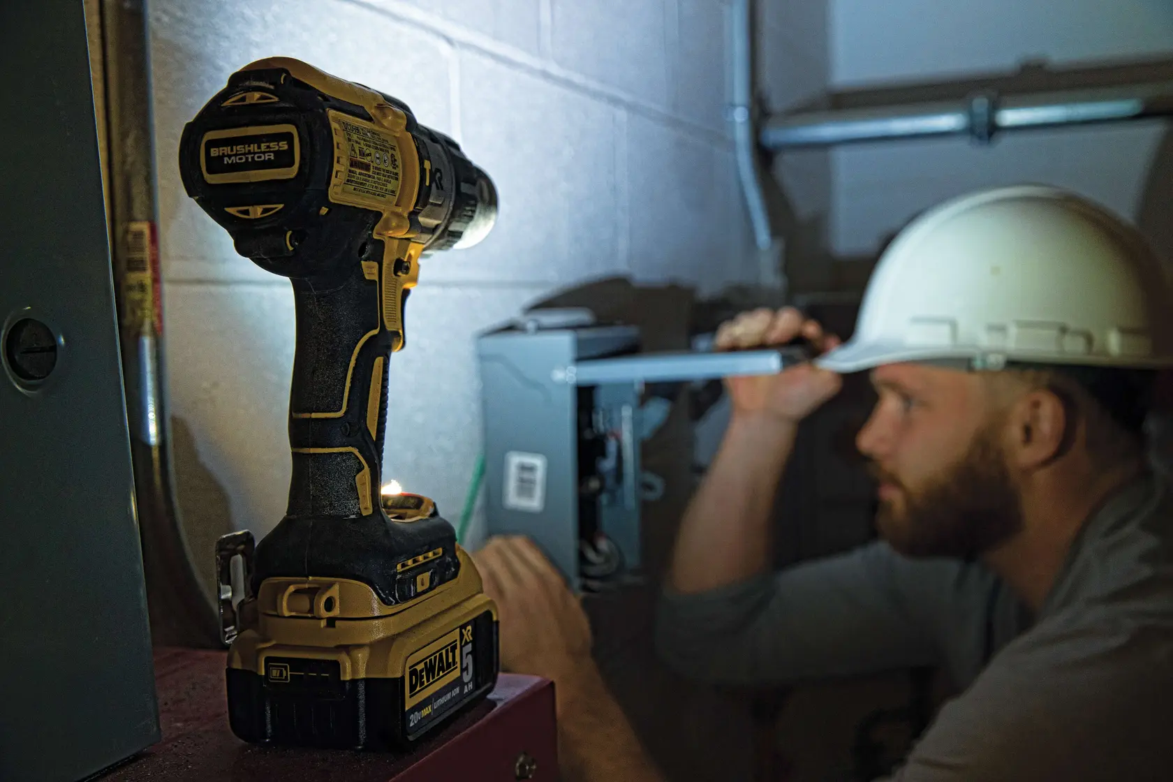 A DEWALT brushless cordless drill is shown resting on a surface in the foreground, with a person wearing a safety helmet working on an electrical box in the background. The person's face is blurred for privacy.