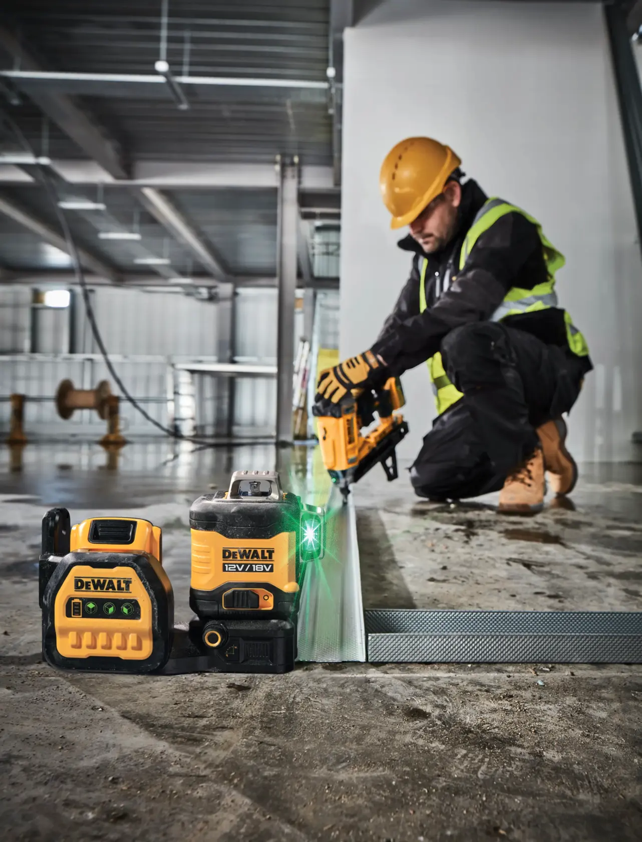 DEWALT laser level and battery on floor; worker uses power tool in construction site background.