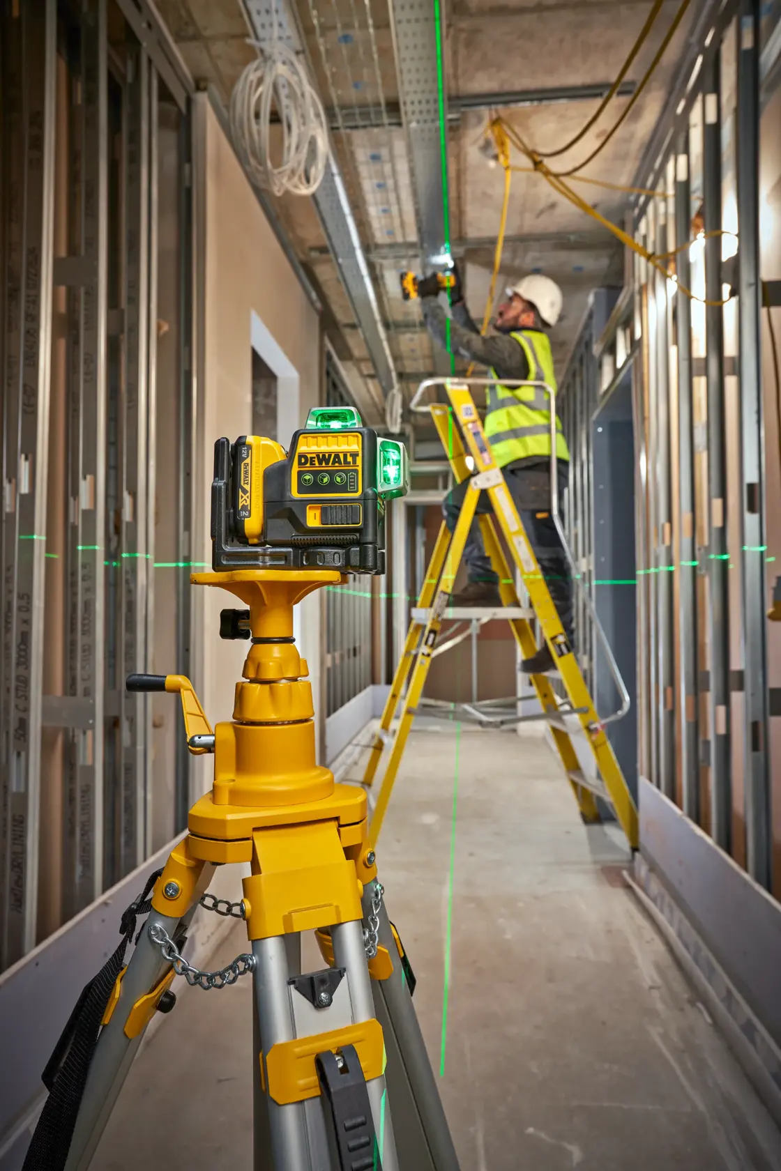 DEWALT laser level on tripod projects green lines in a construction area; worker uses ladder for installation tasks.