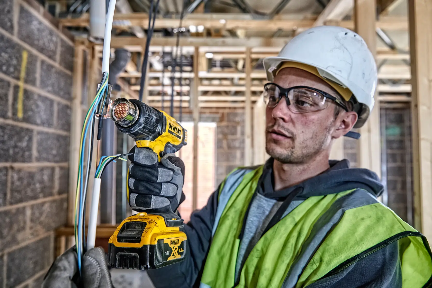 A person wearing safety gear uses a DEWALT DCE530N cordless heat gun to work on electrical wires at a construction site. The individual's face is obscured for privacy.