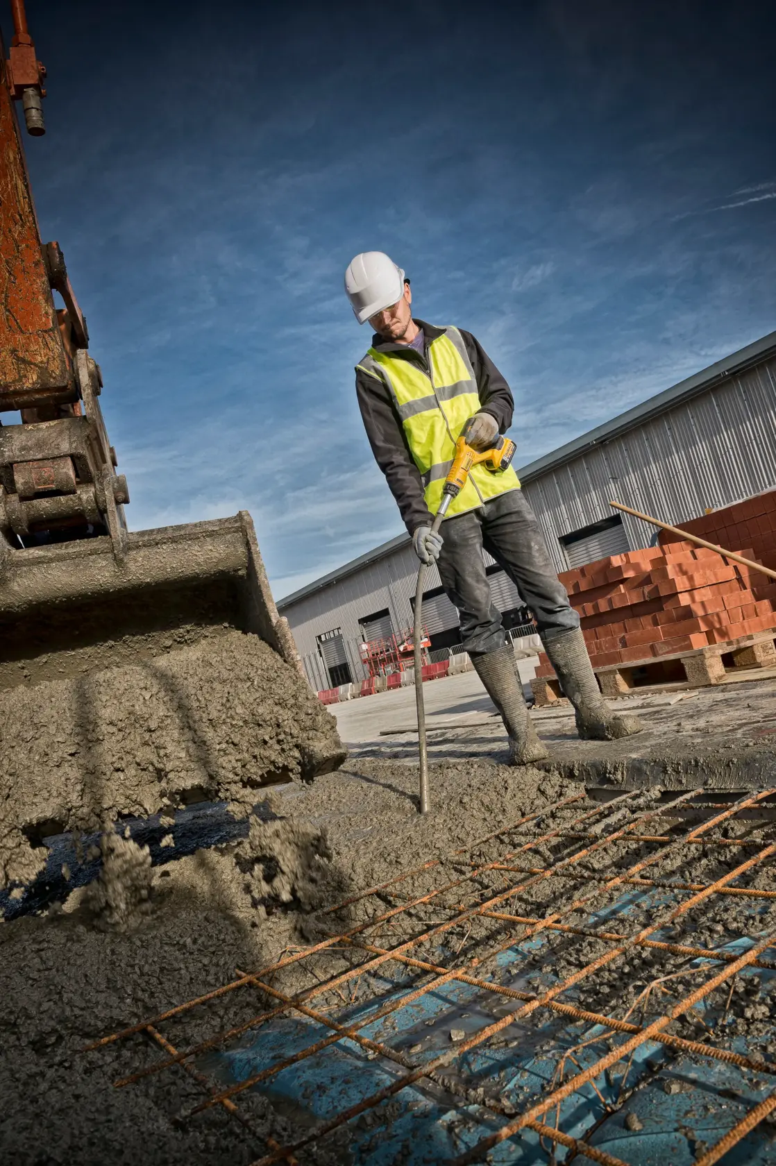 A construction worker wearing a high-visibility vest and helmet uses a DEWALT concrete vibrator on a building site, next to a pouring excavator and steel reinforcement mesh.