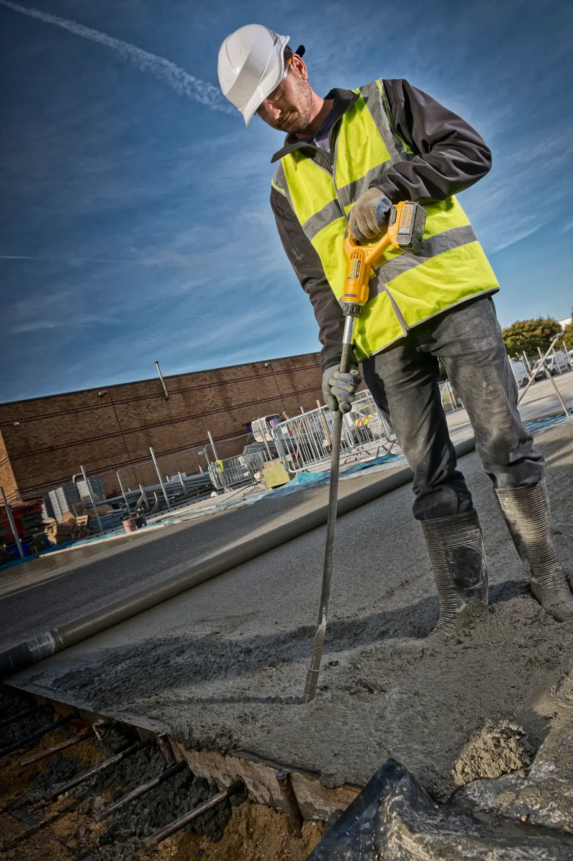A worker wearing a safety helmet, hi-vis vest, gloves, and boots operates a DEWALT DCE531N tool to vibrate freshly poured concrete at a construction site.