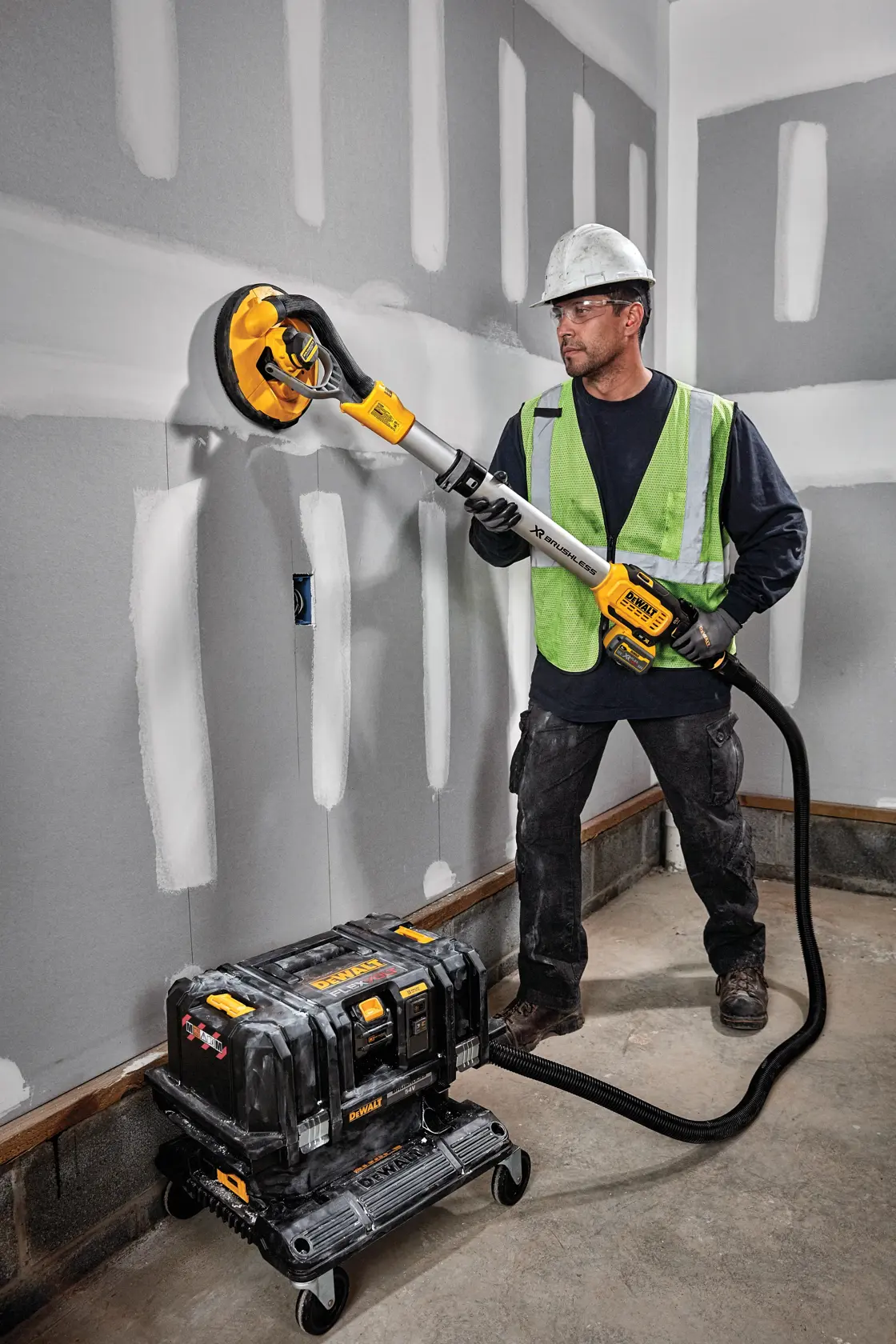 A construction worker wearing a hard hat and safety vest is sanding a drywall using a DEWALT cordless drywall sander, model DCE800T2. The sander is connected by a hose to a portable dust extractor placed on the floor.