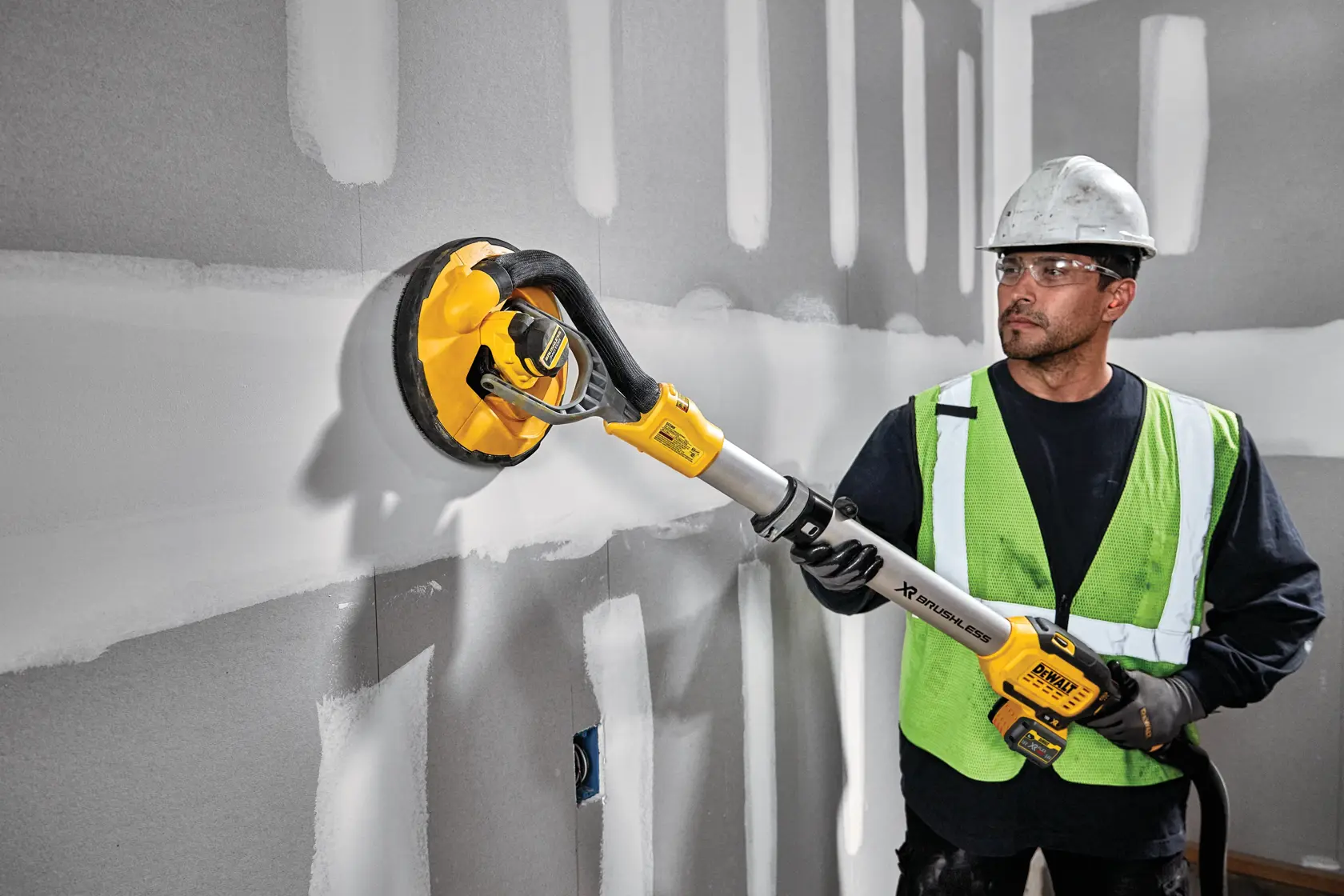 A person wearing a hard hat and a safety vest is using a DEWALT DCE800T2 cordless drywall sander on a wall with unfinished plaster joints.