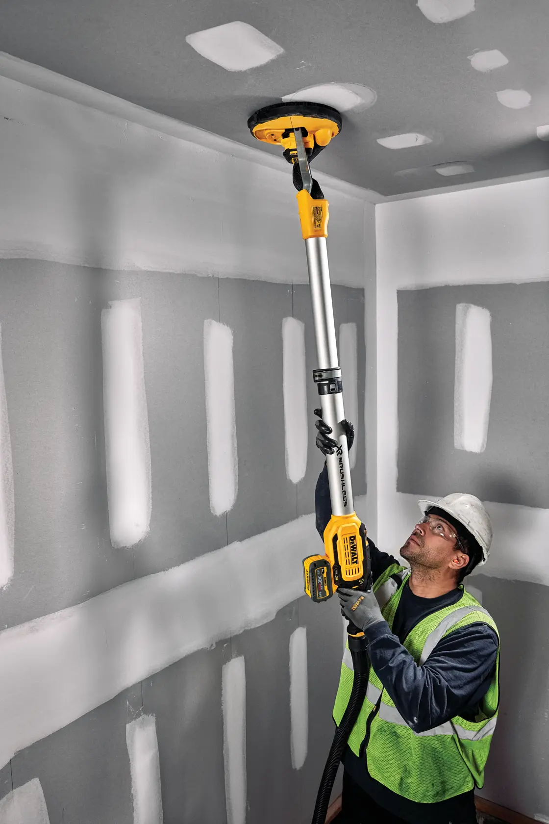 A worker in a safety vest and hard hat uses a DEWALT cordless drywall sander with an extension pole to sand the ceiling in a room with unfinished drywall.
