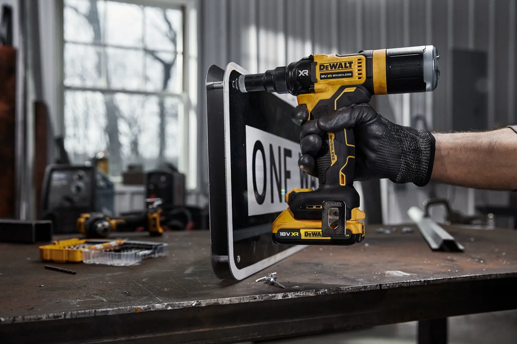 En person med svart arbeidshanske bruker en DEWALT DCF403NT 18V XR batteridrill for å bore i et metallskilt på en arbeidsbenk i et verksted.