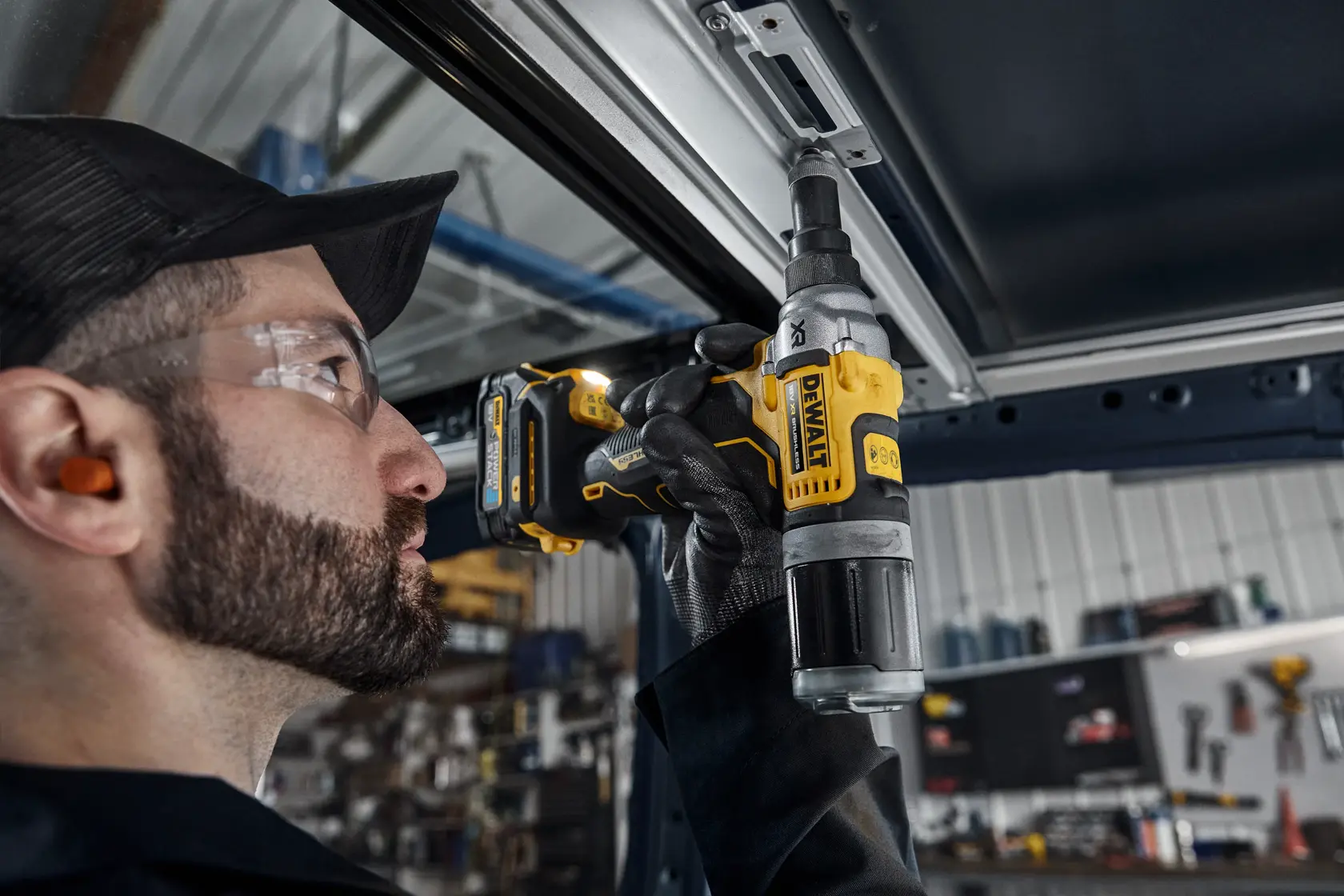 En person med hørselvern og hansker bruker et trådløst DEWALT DCF414N elverktøy for å feste eller løsne en metallkomponent innendørs i et industri- eller verkstedmiljø.