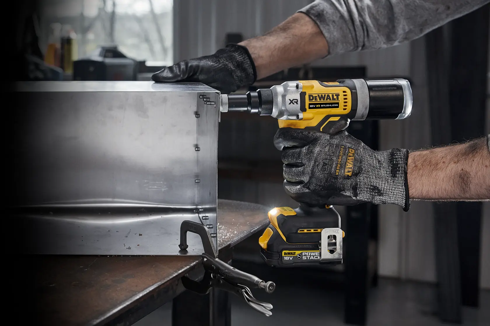 En person med arbeidshansker bruker en DEWALT DCF414NT 18V trådløs popnagletang for å feste en metallplate i et industriverksted.