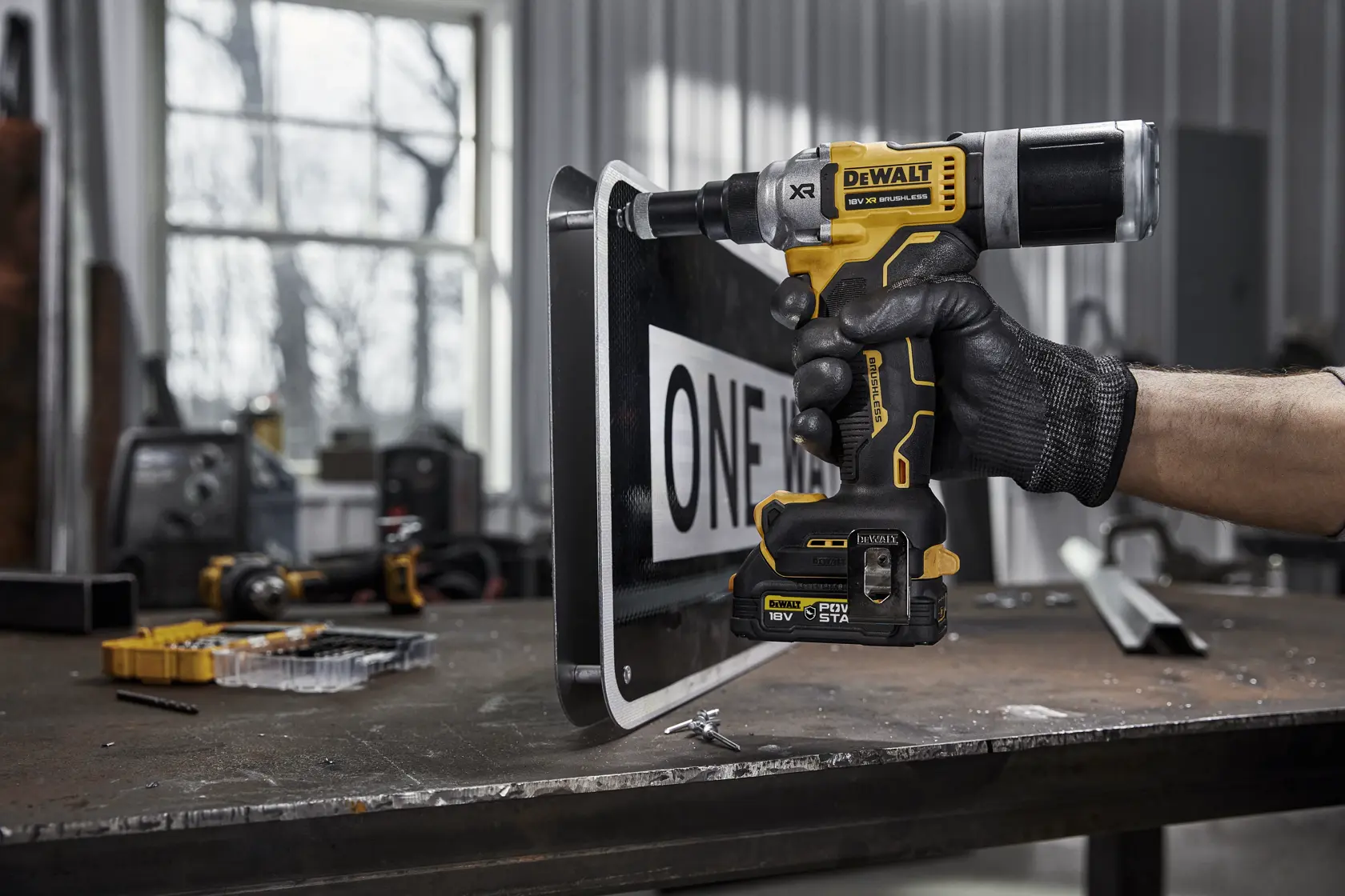 En person med svart hanske bruker et DEWALT DCF414NT trådløst elektrisk verktøy på et metallskilt i et verksted. Verktøyet har gul og svart DEWALT-merking og er plassert mot kanten av skiltet. Ulike verktøy og deler er synlige på arbeidsbenken i bakgrunnen.
