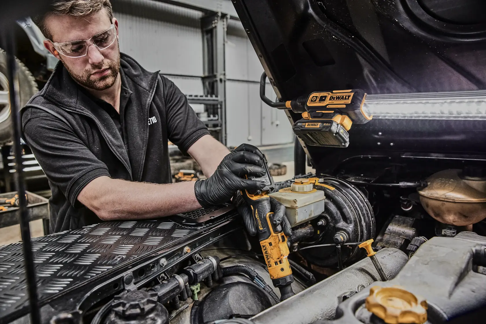 A person wearing black gloves uses a DEWALT DCF503L1G cordless ratchet tool to work on the engine of a vehicle. The vehicle hood is open, and a DEWALT work light is attached to the hood to illuminate the workspace. The person's face is blurred for privacy.