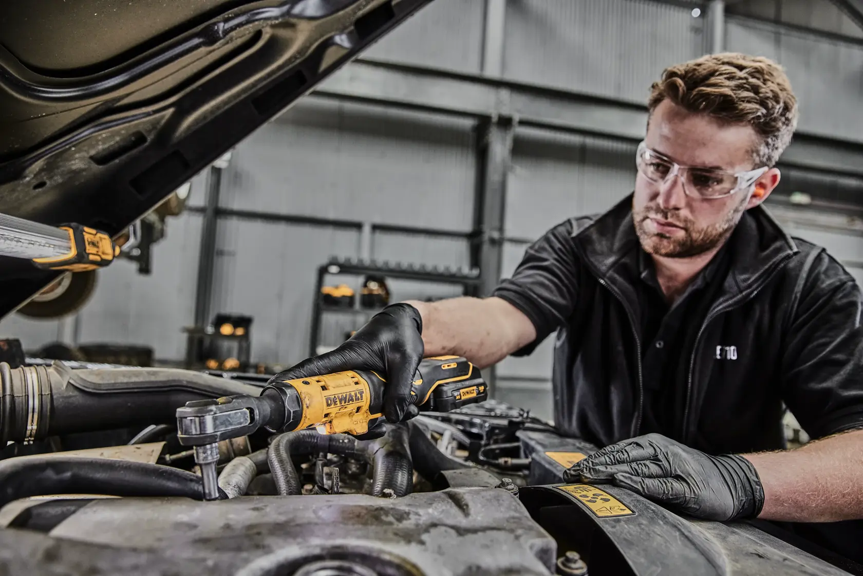 A person wearing black gloves is using a DEWALT DCF504N cordless ratchet tool to work on a car engine in an industrial garage. The face of the person is blurred for privacy.