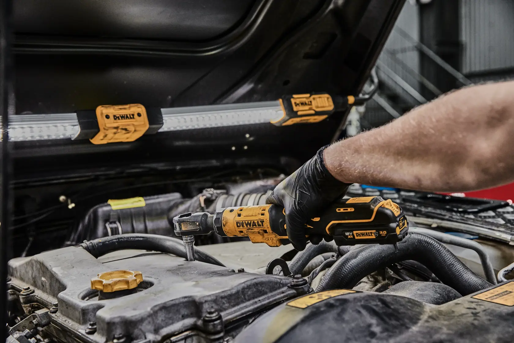 A person uses a DEWALT cordless ratchet tool (model DCF504N) to work on a car engine in a garage setting. An LED work light with DEWALT branding is attached to the car hood for illumination.