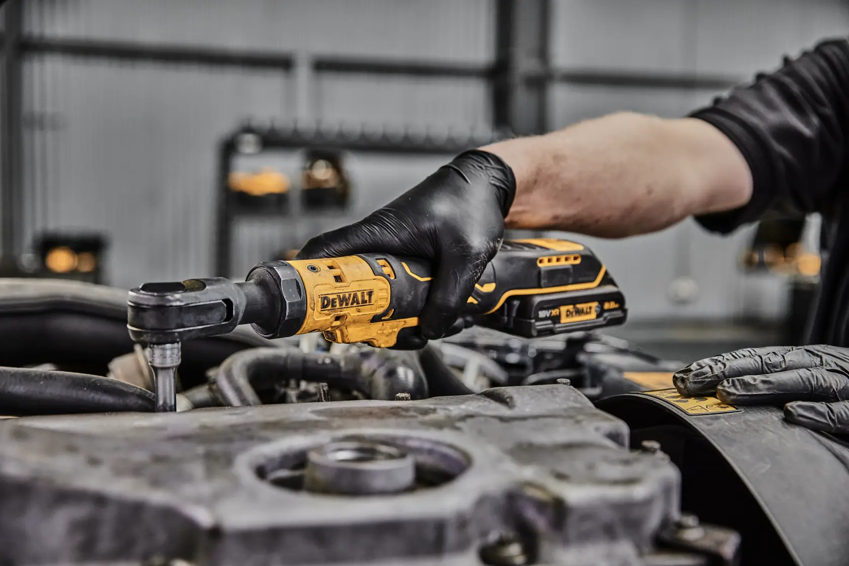 A person wearing black gloves uses a DEWALT cordless ratchet wrench to work on an engine part in a workshop.