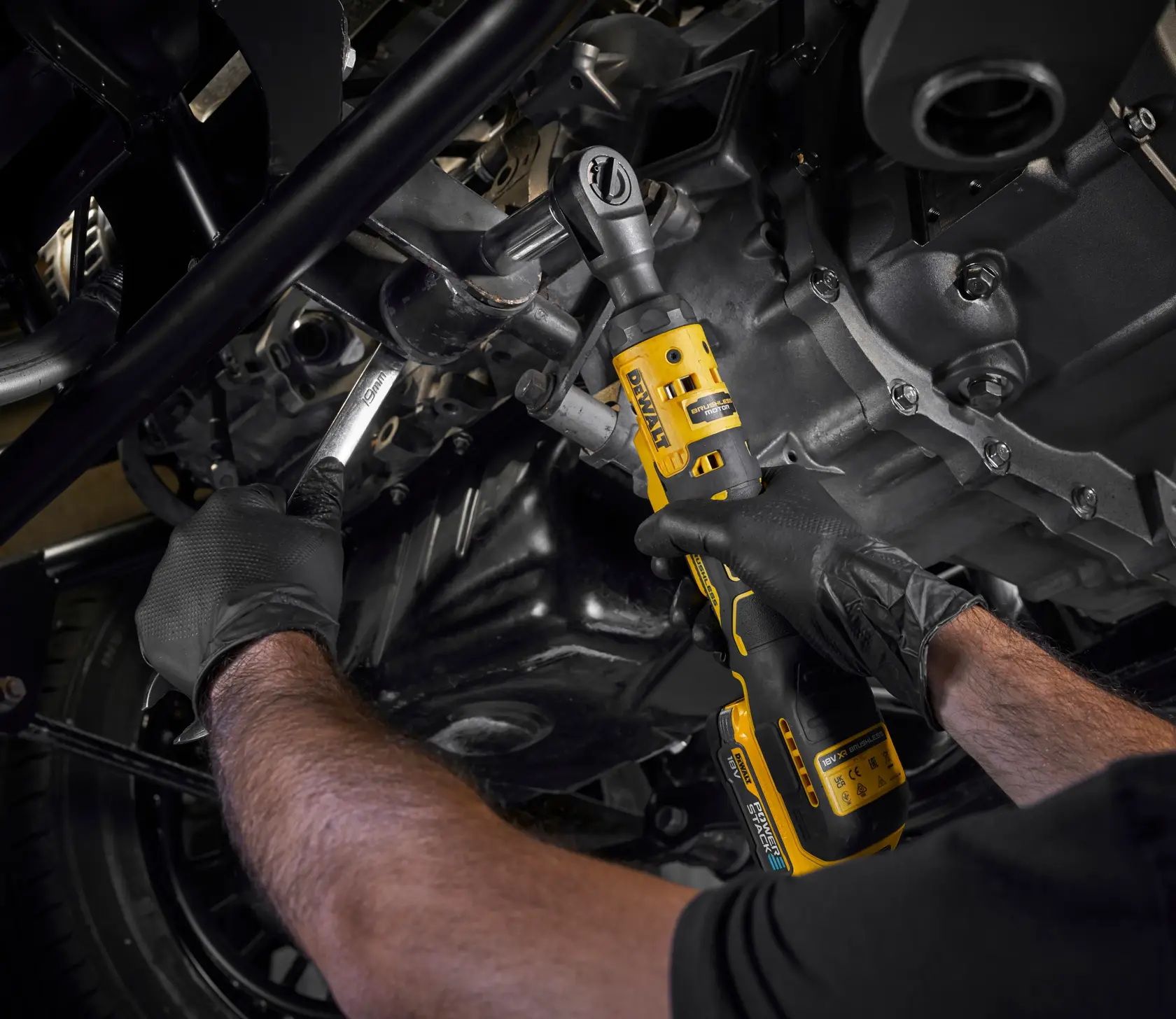 Person uses a DEWALT cordless ratchet tool to work on a vehicle engine, wearing black gloves.