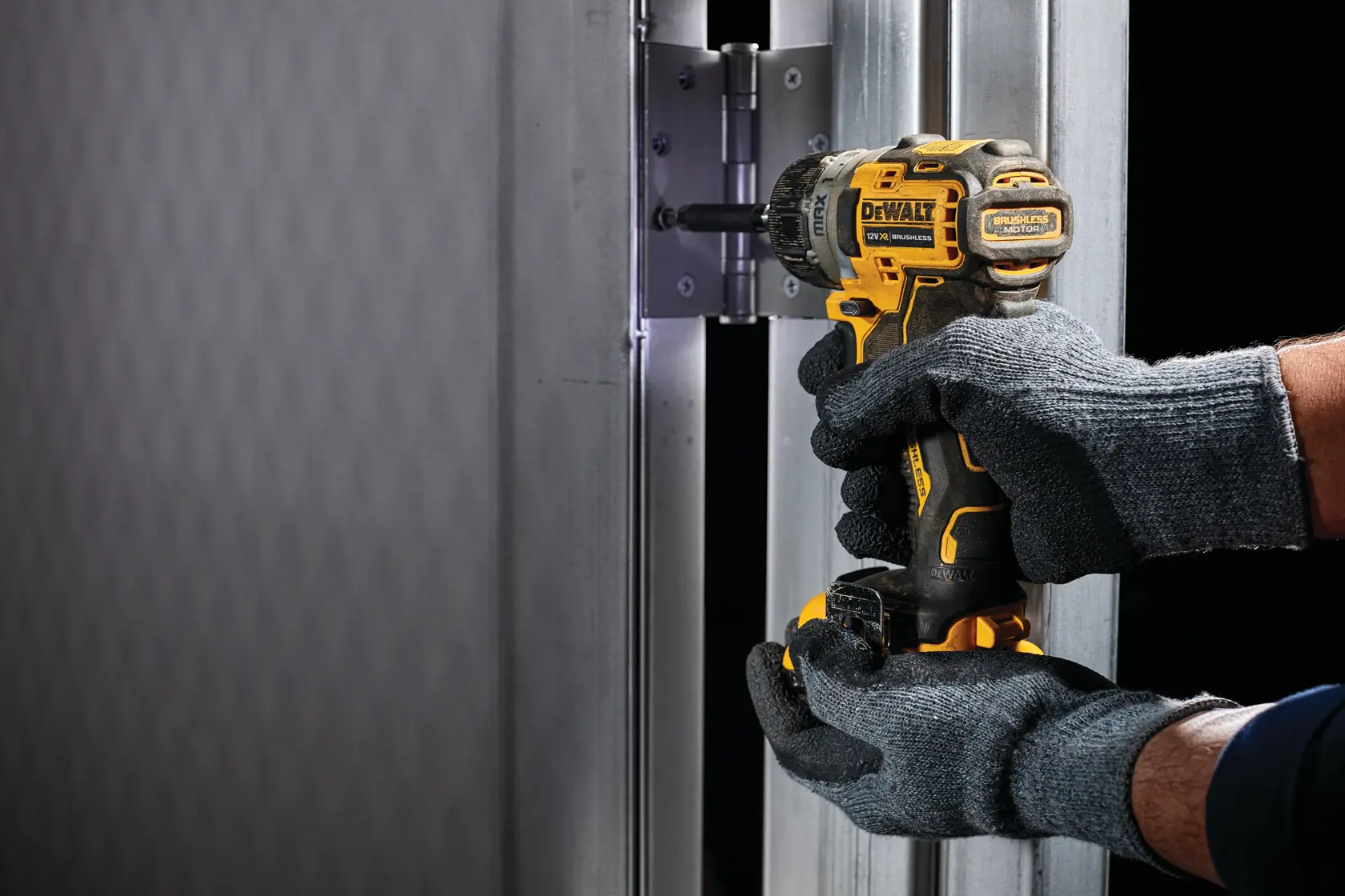Gloved hands use a DEWALT DCF601N cordless screwdriver to tighten screws on a metal hinge.