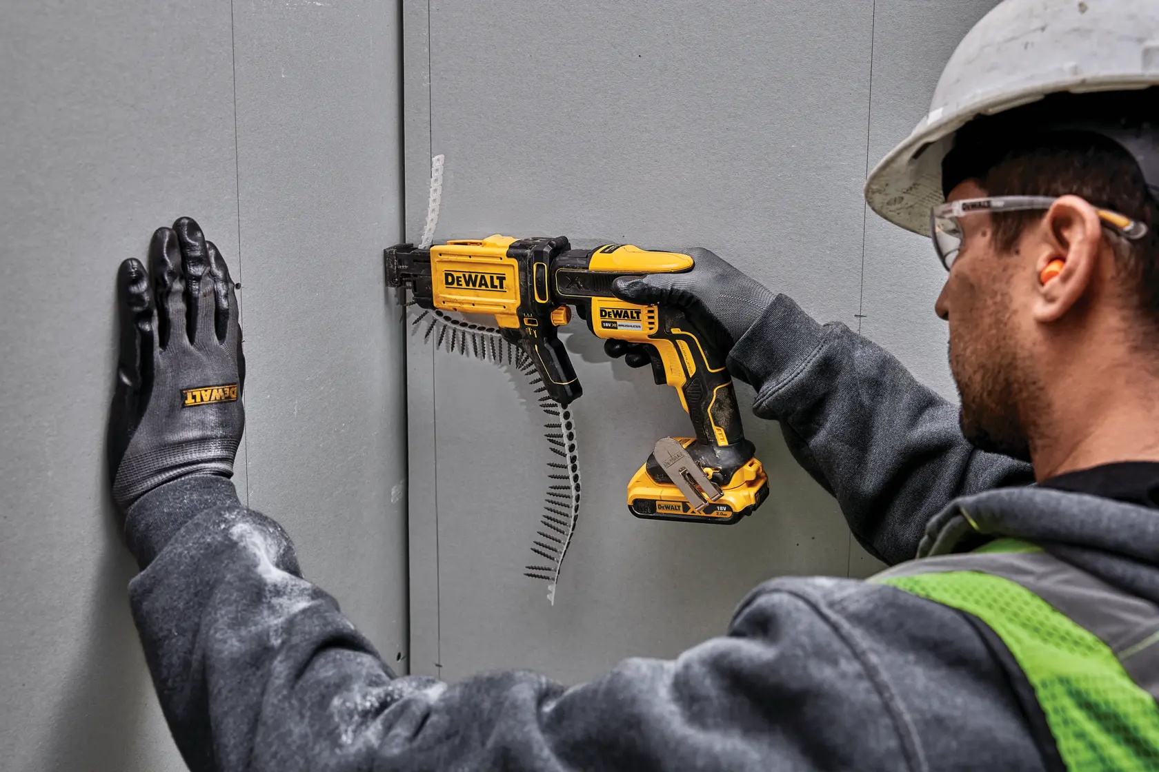 Worker uses DEWALT DCF6202 tool to attach drywall, wearing gloves, helmet, and safety glasses.