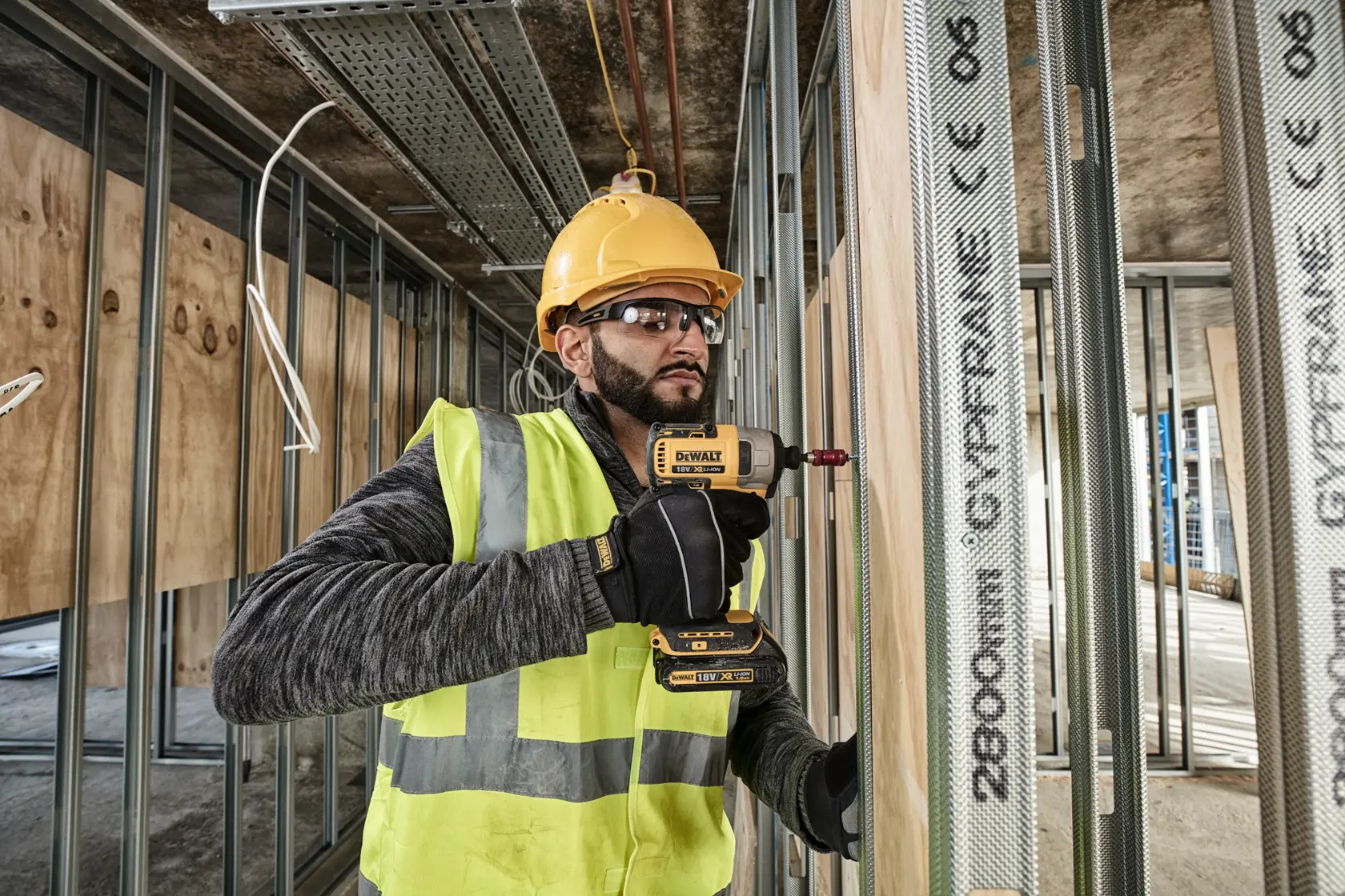 Worker using DEWALT DCF787N drill on metal studs at construction site, wearing safety gear and yellow helmet.
