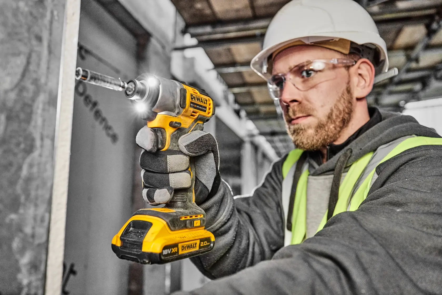 Persona con casco di sicurezza e giubbotto ad alta visibilità che utilizza un avvitatore a impulsi cordless DEWALT DCF840D2T in un cantiere. Il volto della persona è sfocato per garantire la privacy.