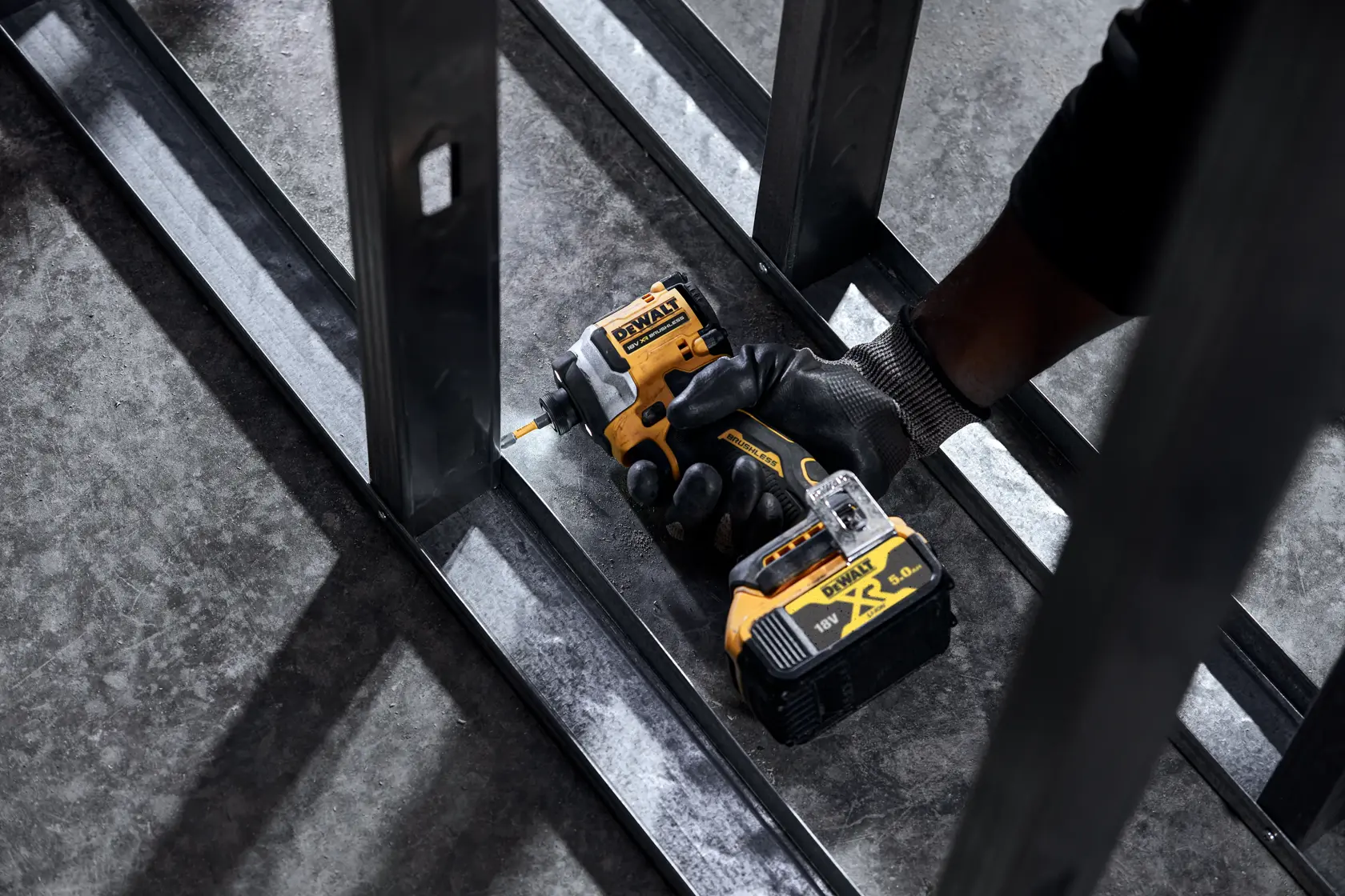 Gloved hand using a DEWALT cordless impact driver on a metal frame surface.