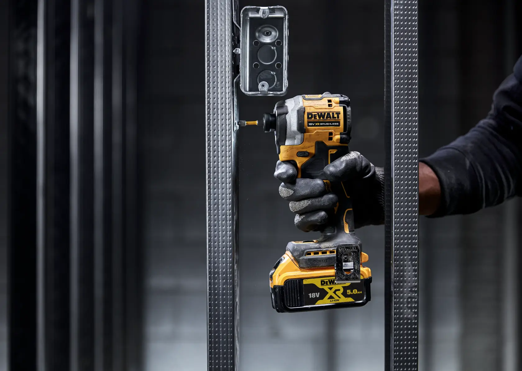 Gloved hand using a DEWALT DCF850NT impact driver to screw into a metal frame near an electrical box.