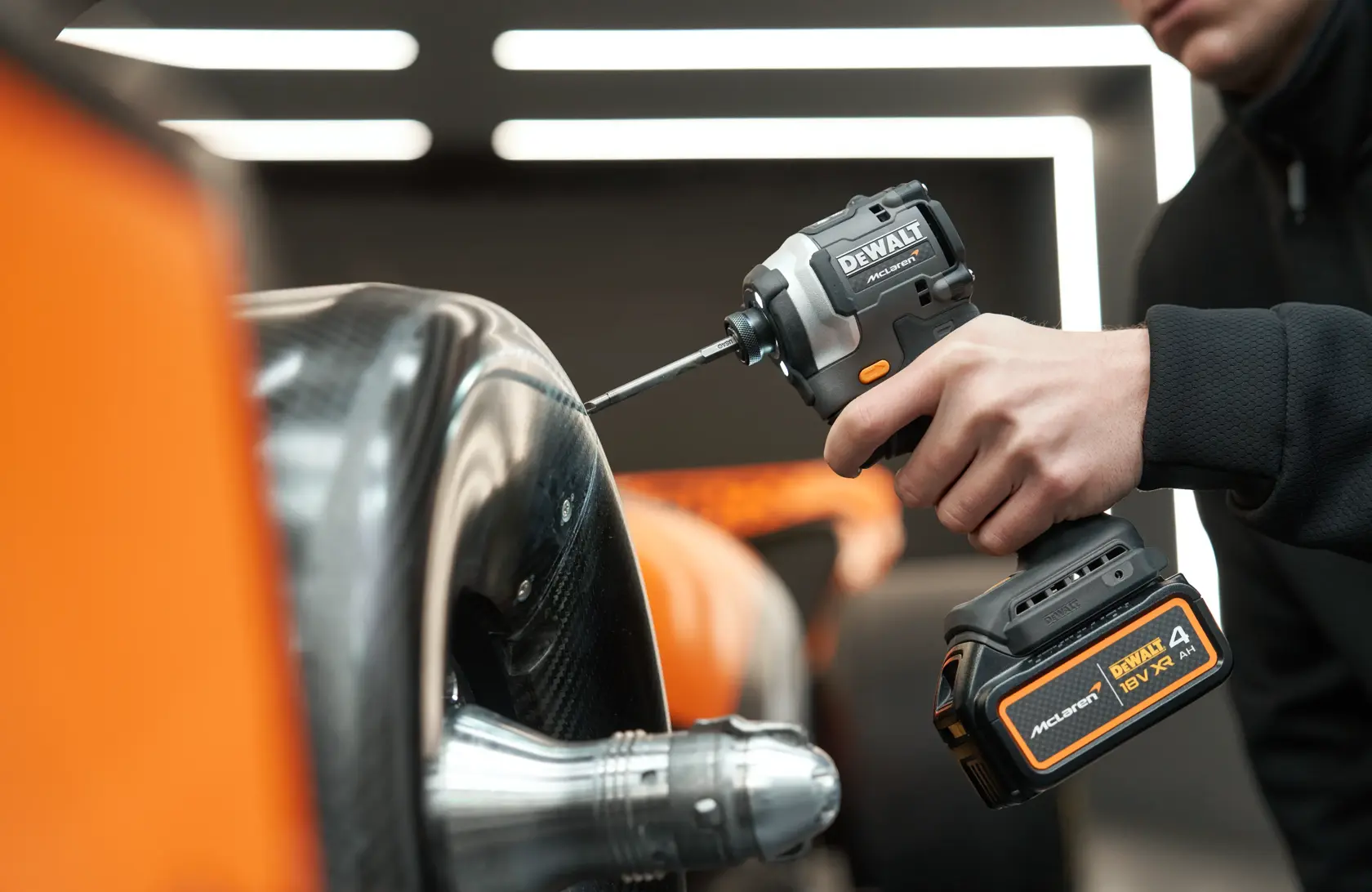Person uses a DEWALT cordless drill on a carbon fiber vehicle part indoors. McLaren branding visible.