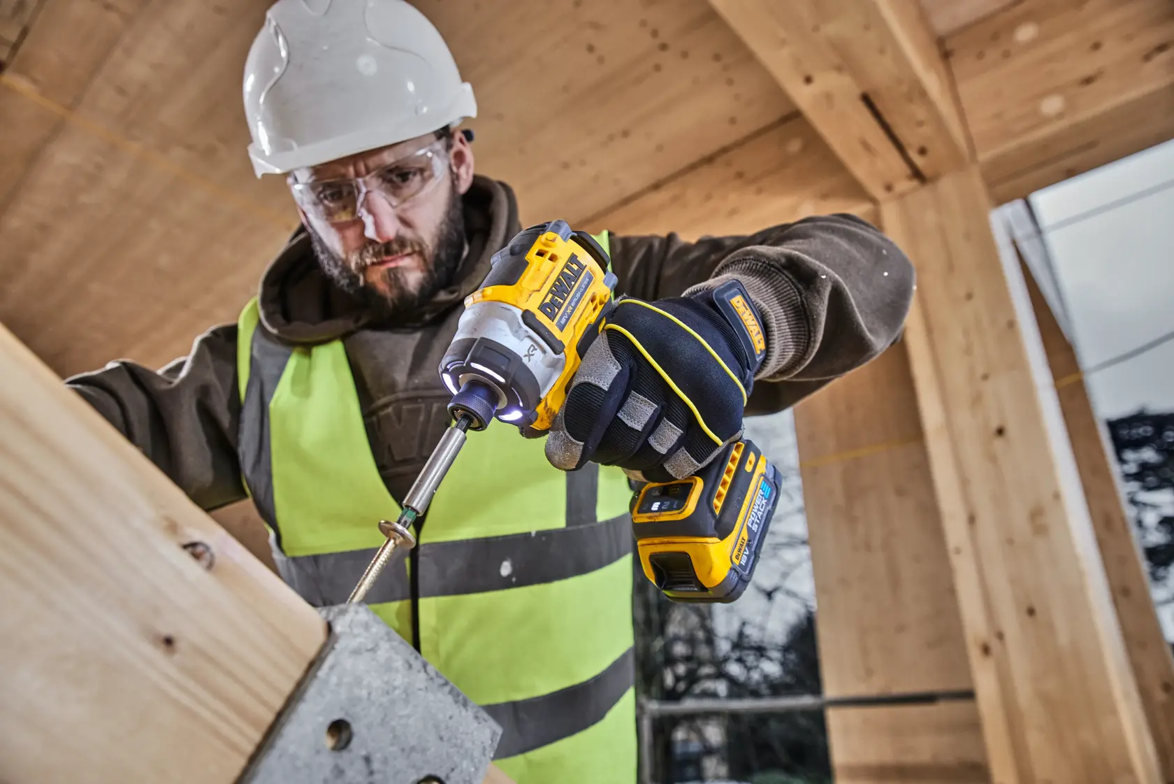 Worker uses DEWALT DCF860E2T impact driver with gloves and helmet on a wooden construction site.