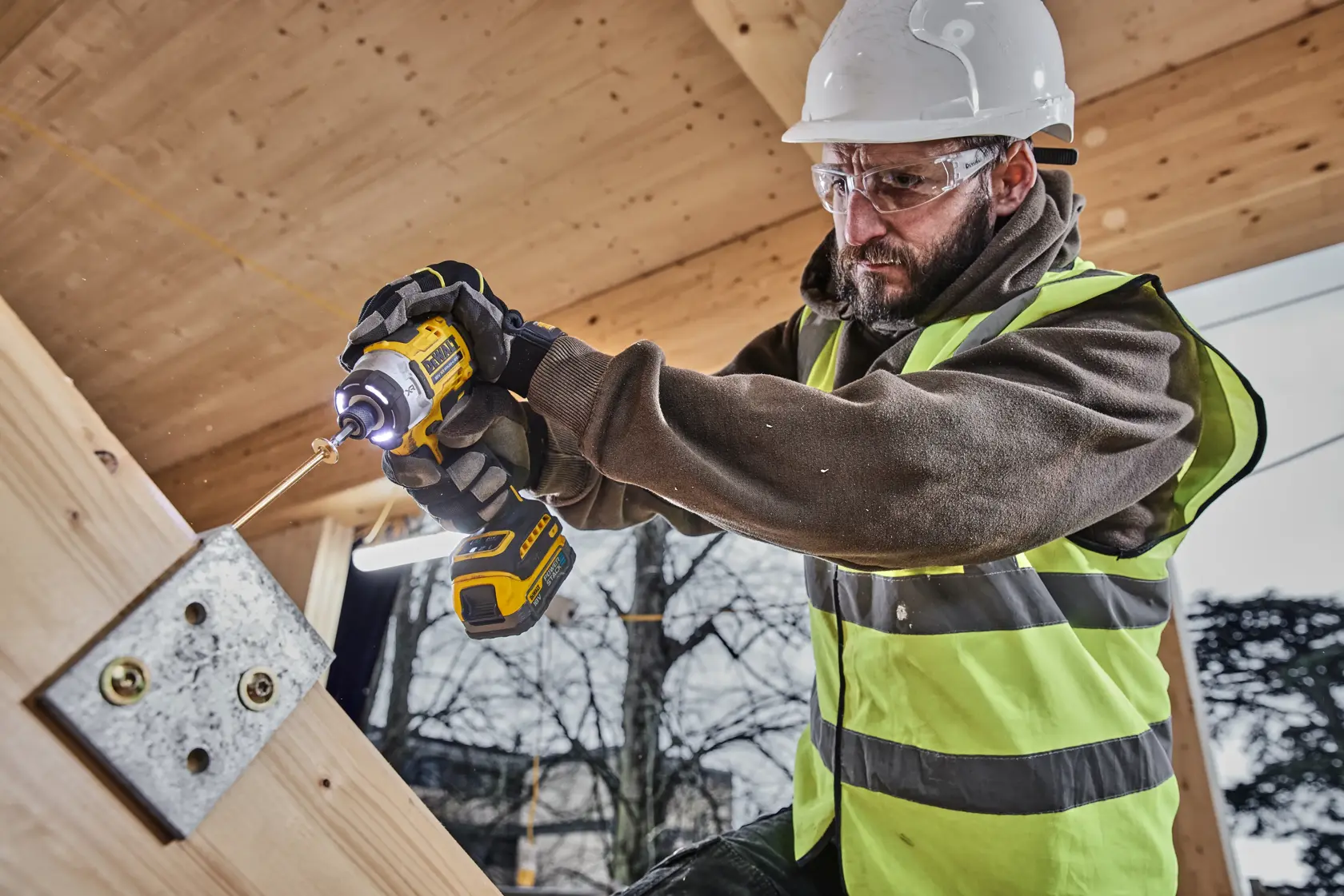 Worker in safety gear uses a DEWALT power tool to drill into wood at a construction site.