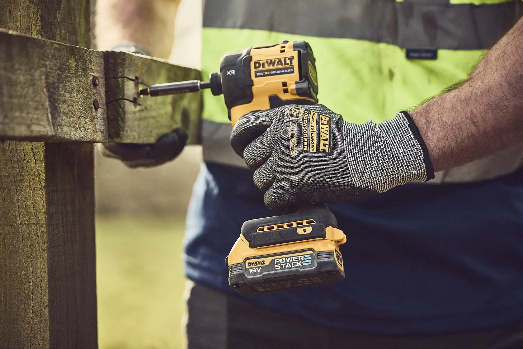 Gloved hand using DEWALT cordless impact driver to fasten screw into wooden post outdoors.