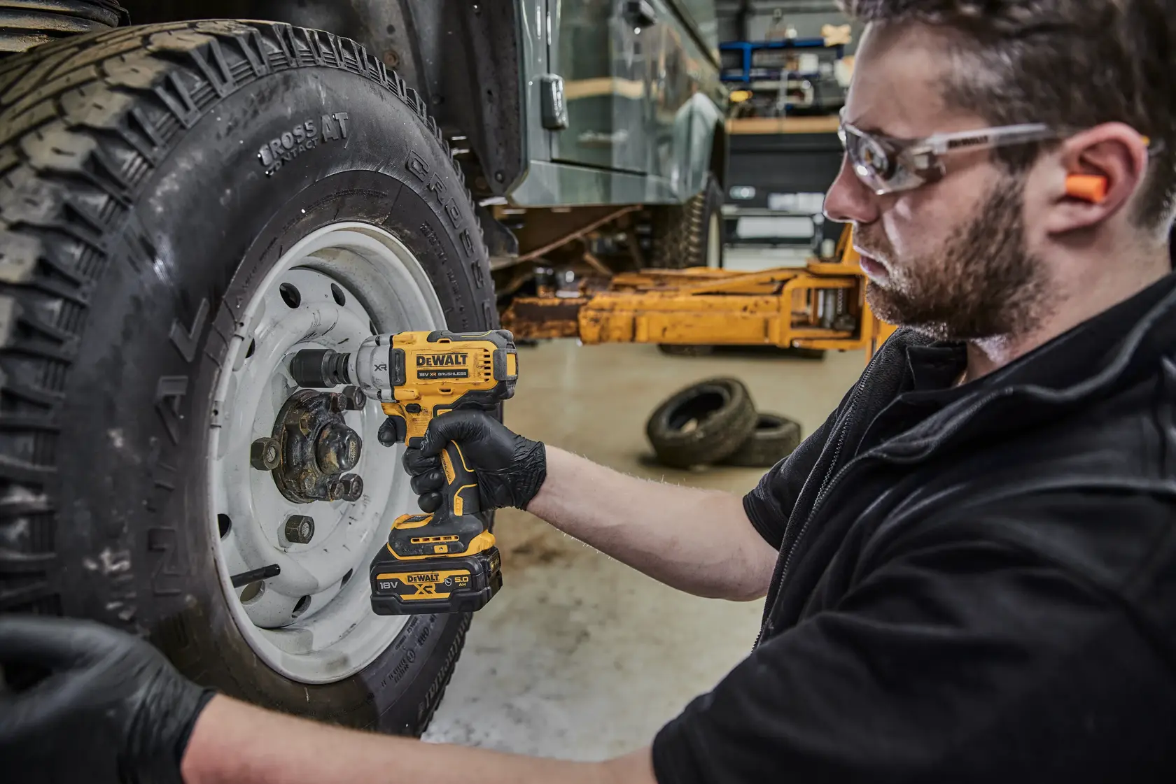 A person wearing gloves uses a DEWALT cordless impact wrench (model DCF891P2G) to work on the lug nuts of a vehicle tire in a garage setting. The person's face is blurred for privacy.