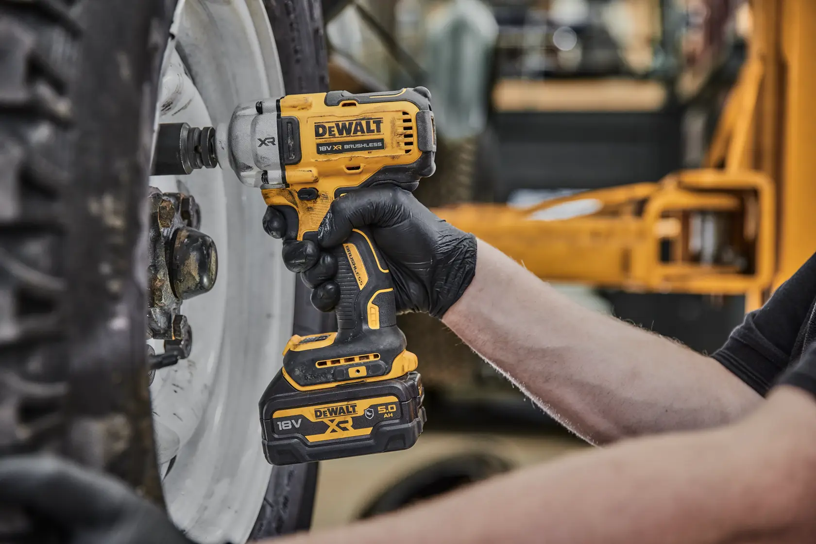A person wearing black gloves is using a yellow DEWALT cordless impact wrench (model DCF891P2G, 18V XR) to work on the bolts of a large vehicle wheel.