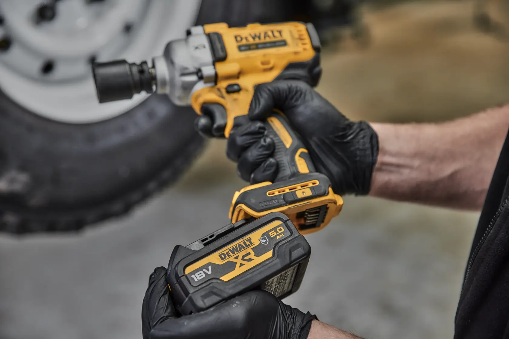 A person wearing black gloves is holding a yellow and black DEWALT cordless impact wrench and a DEWALT 18V XR 5.0Ah lithium-ion battery. The background shows a blurred tire.