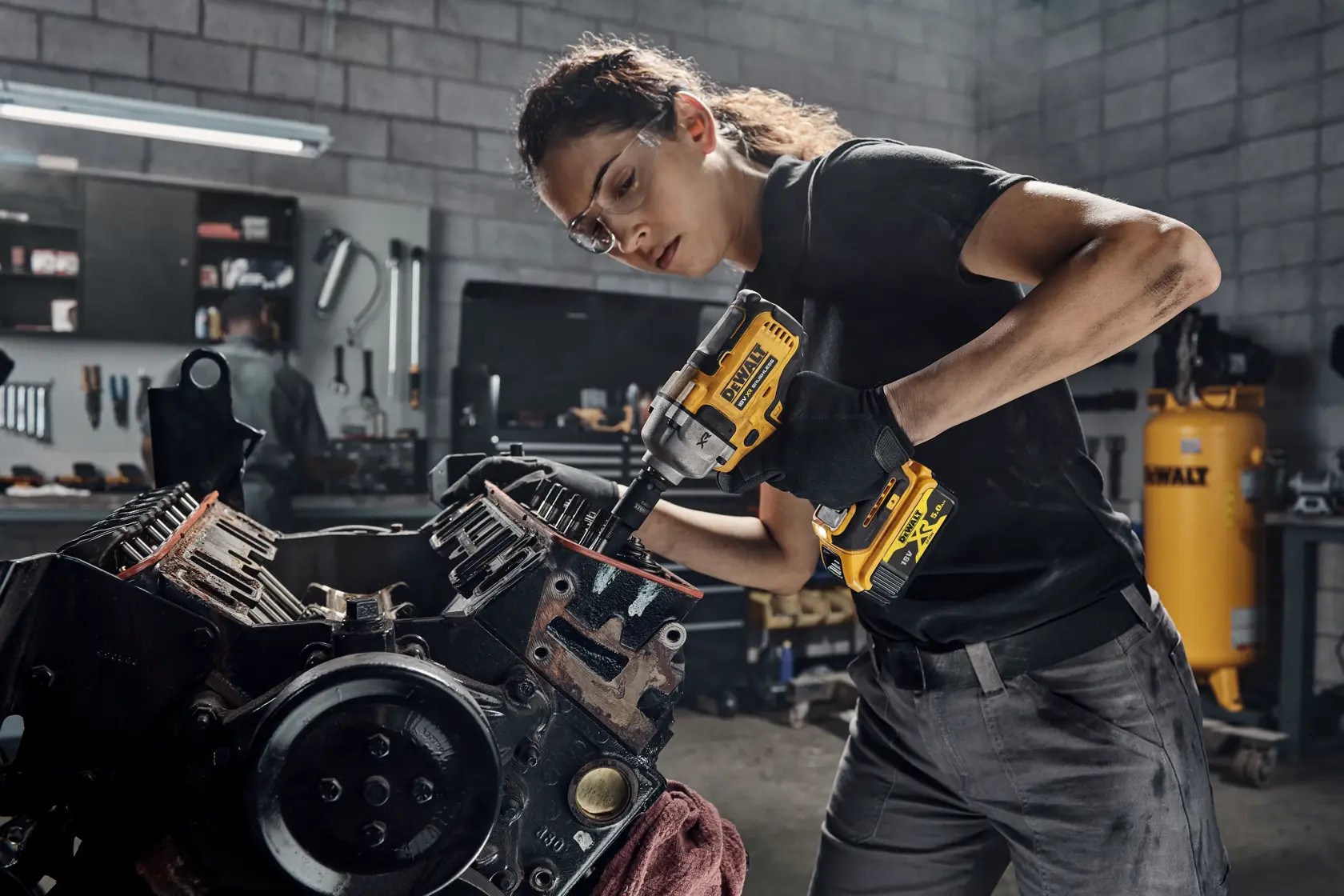 Person uses a DEWALT DCF891P2T impact wrench on a machine part in a workshop with tools in the background.