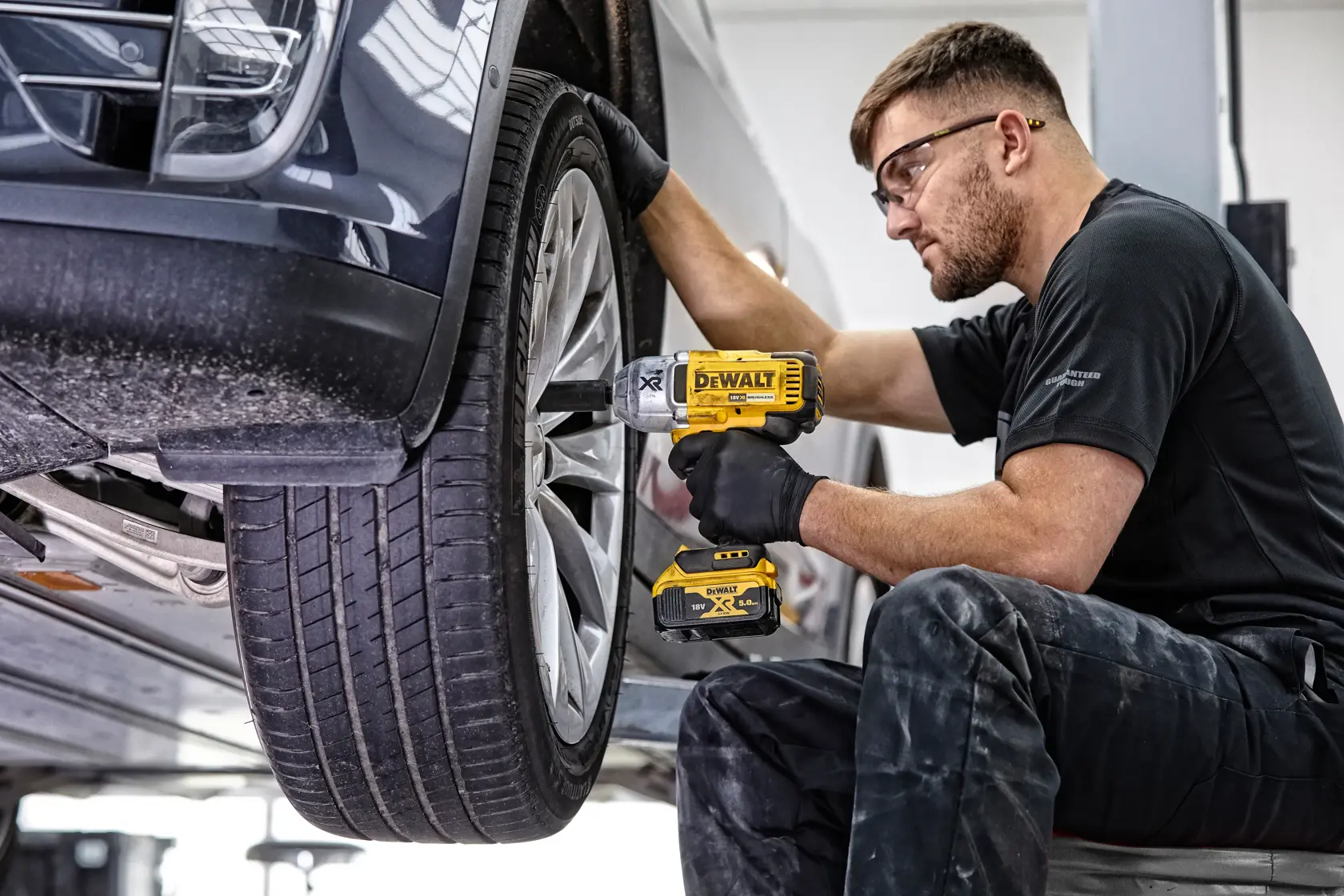 Person uses DEWALT impact wrench on car wheel in garage, wearing gloves and safety glasses.