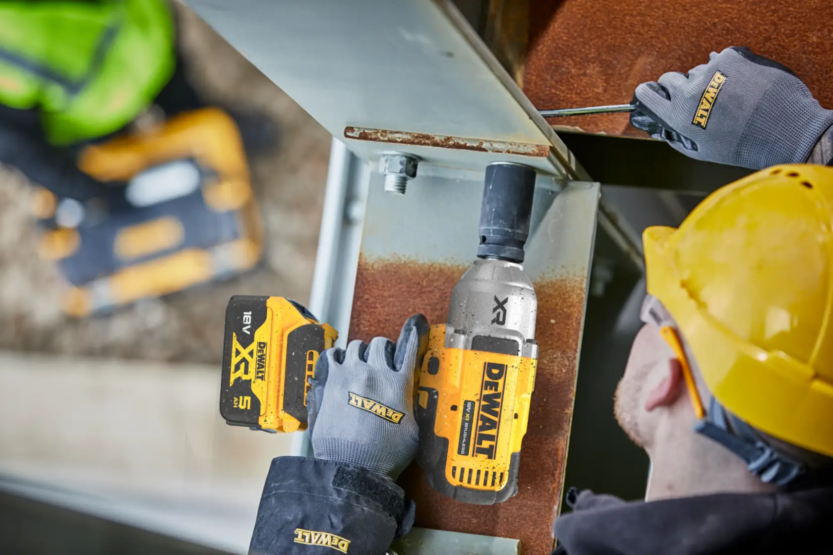 Worker uses DEWALT cordless impact wrench on metal beam while wearing gloves and yellow helmet.