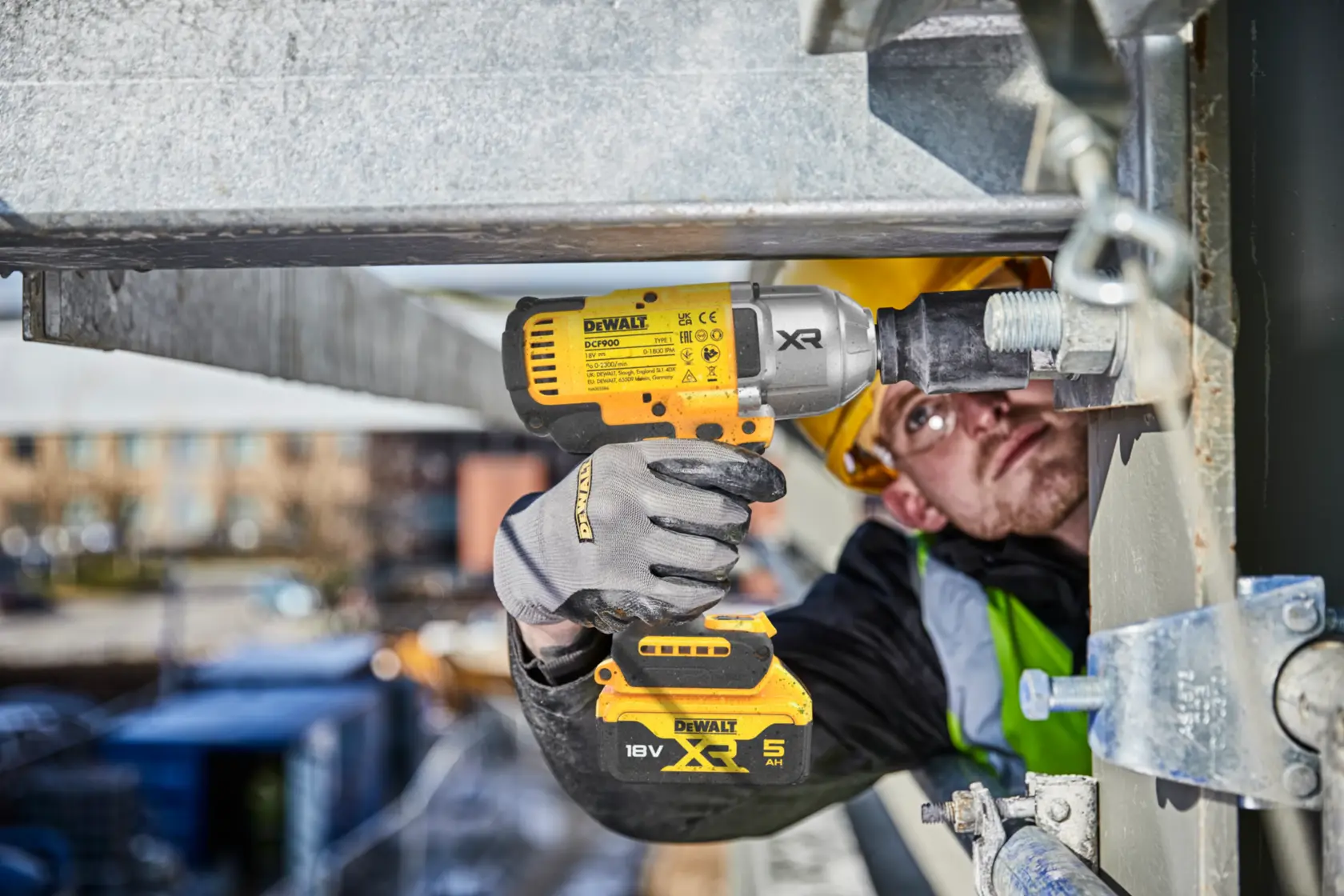 Worker uses DEWALT DCF900P2T impact wrench on metal structure outdoors. Battery pack and gloves visible.