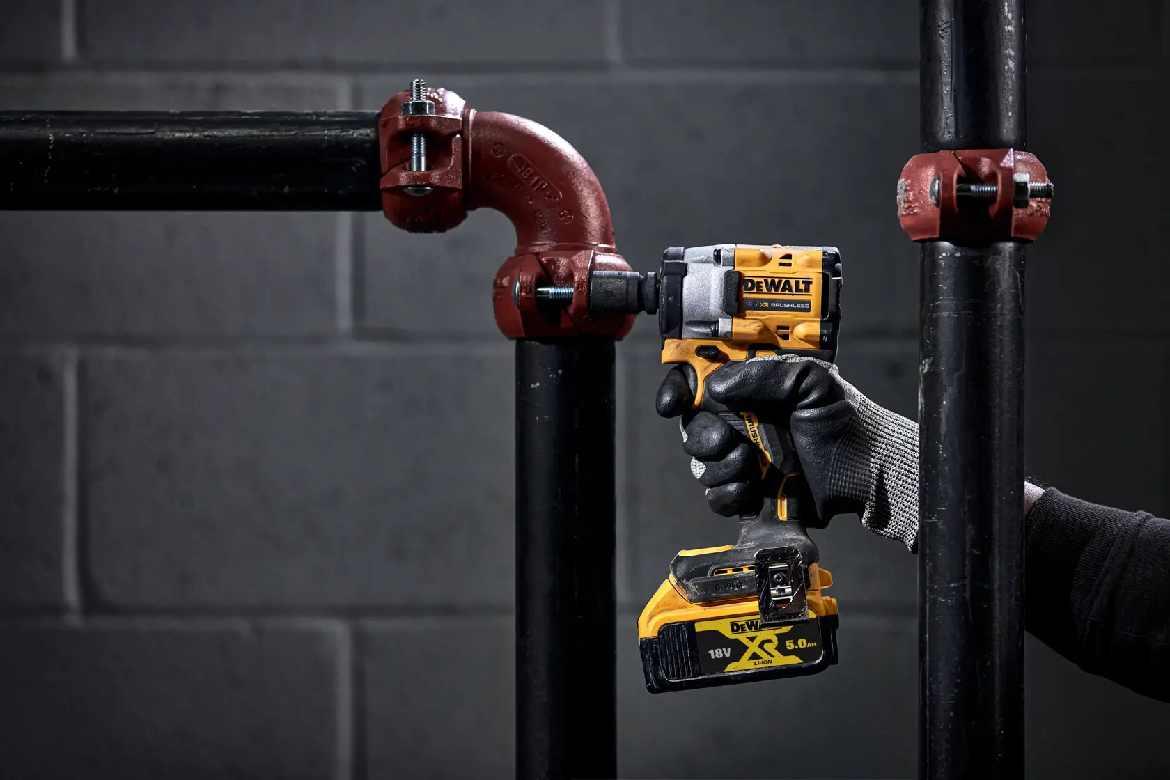 Una persona con i guanti utilizza un avvitatore a impulsi senza fili DEWALT per serrare un bullone su un raccordo per tubi in metallo. L'utensile è giallo e nero e la scena è ambientata in un contesto industriale scuro.