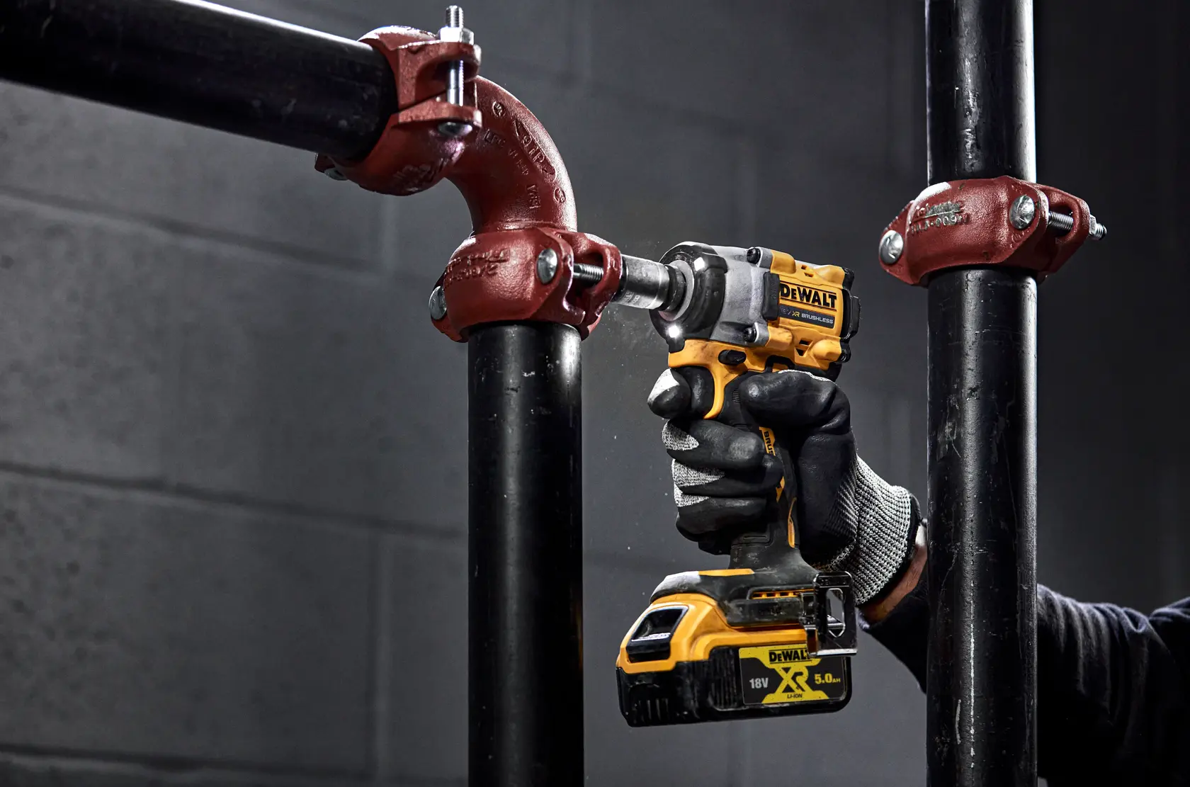 Una persona con guanti da lavoro utilizza un avvitatore a impulsi a batteria giallo e nero DEWALT per stringere un bullone su un tubo metallico nero con raccordo rosso.