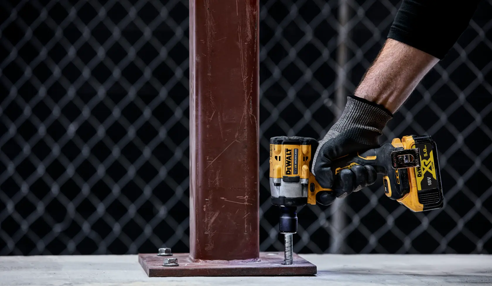 Una persona con un guanto utilizza un avvitatore a impulsi a batteria DEWALT per serrare un bullone su una piastra di base in metallo fissata a una trave d'acciaio verticale.