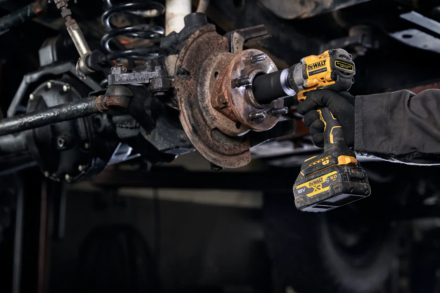 A DEWALT DCF923P2G cordless impact wrench is being used to work on a rusty wheel hub assembly under a vehicle. The tool is held by a gloved hand.