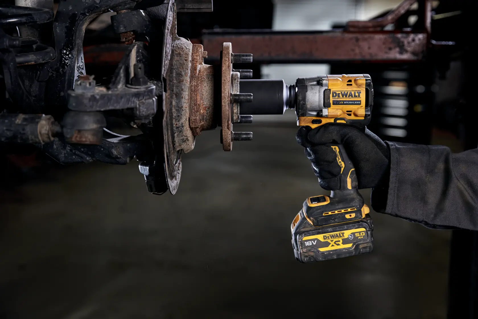 A person using a DEWALT cordless impact wrench (model DCF923P2G) to work on a rusty vehicle wheel hub in a garage or workshop.