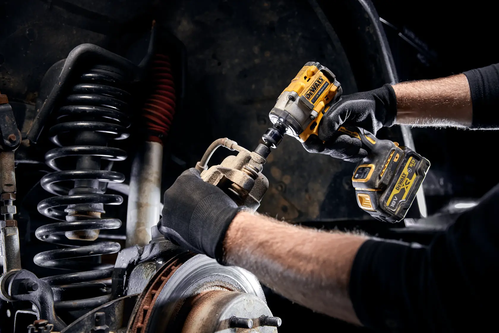A person wearing black gloves is using a yellow DEWALT cordless impact wrench to work on the suspension and brake system of a vehicle.
