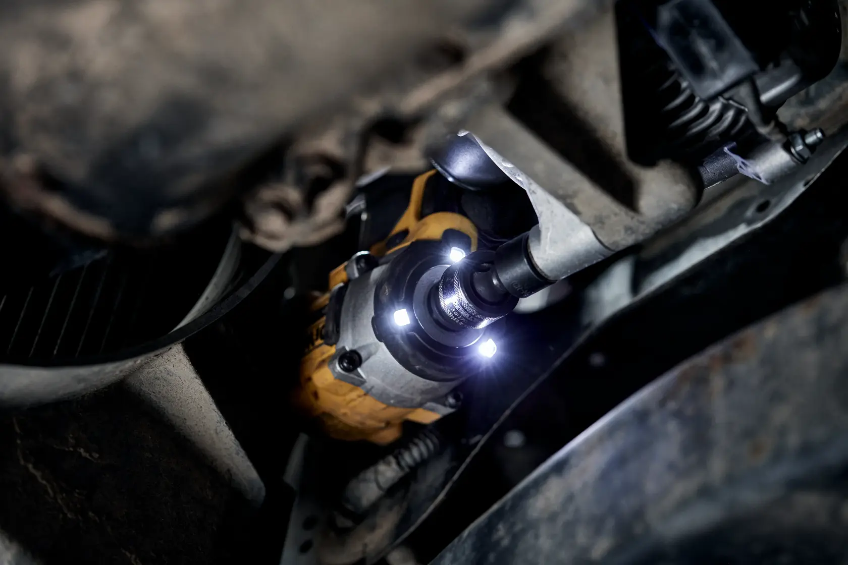 A DEWALT DCF923P2G cordless impact wrench in use, tightening or loosening a bolt in a vehicle's suspension area, illuminated by built-in LED lights.