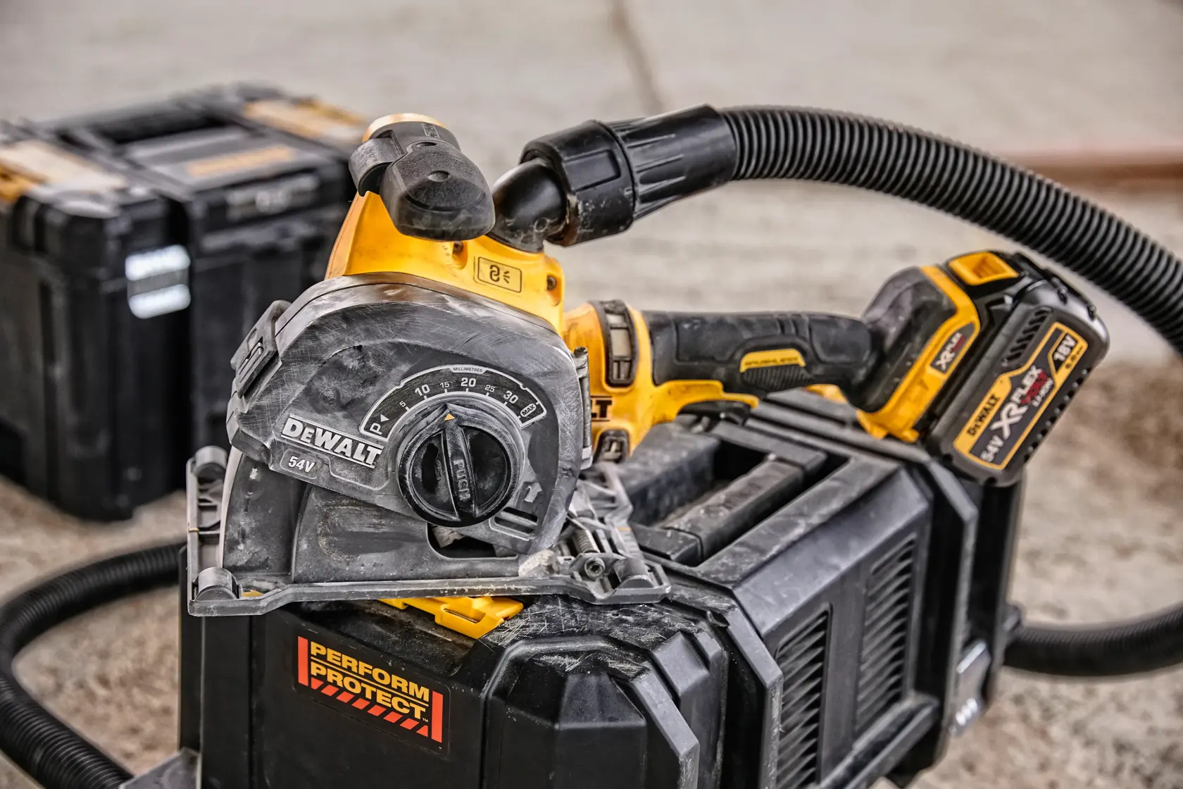 A close-up image of a DEWALT cordless power tool, specifically a cutting tool with dust extraction hose attached, resting on a black tool storage box at a workshop. Another toolbox is visible in the background.