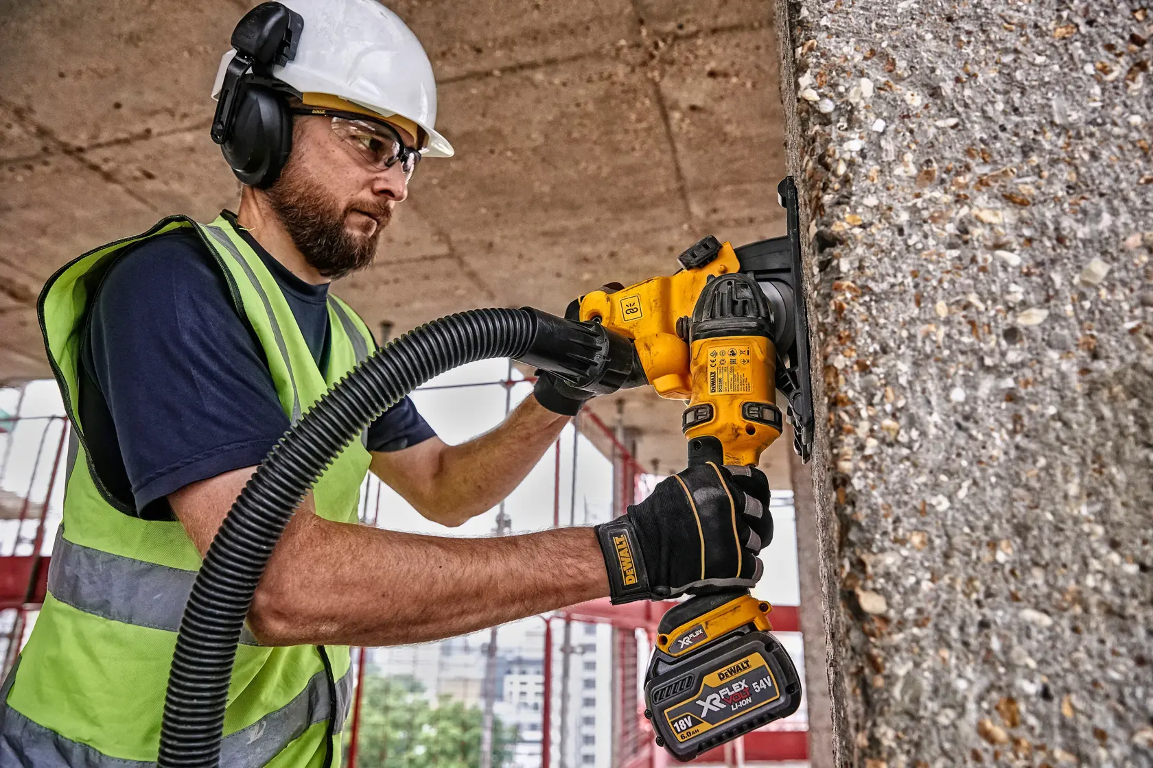 A worker wearing a safety helmet, ear protection, and high-visibility vest uses a DEWALT DCG200T2 cordless power tool for grinding concrete on a construction site. The worker's face is blurred for privacy.