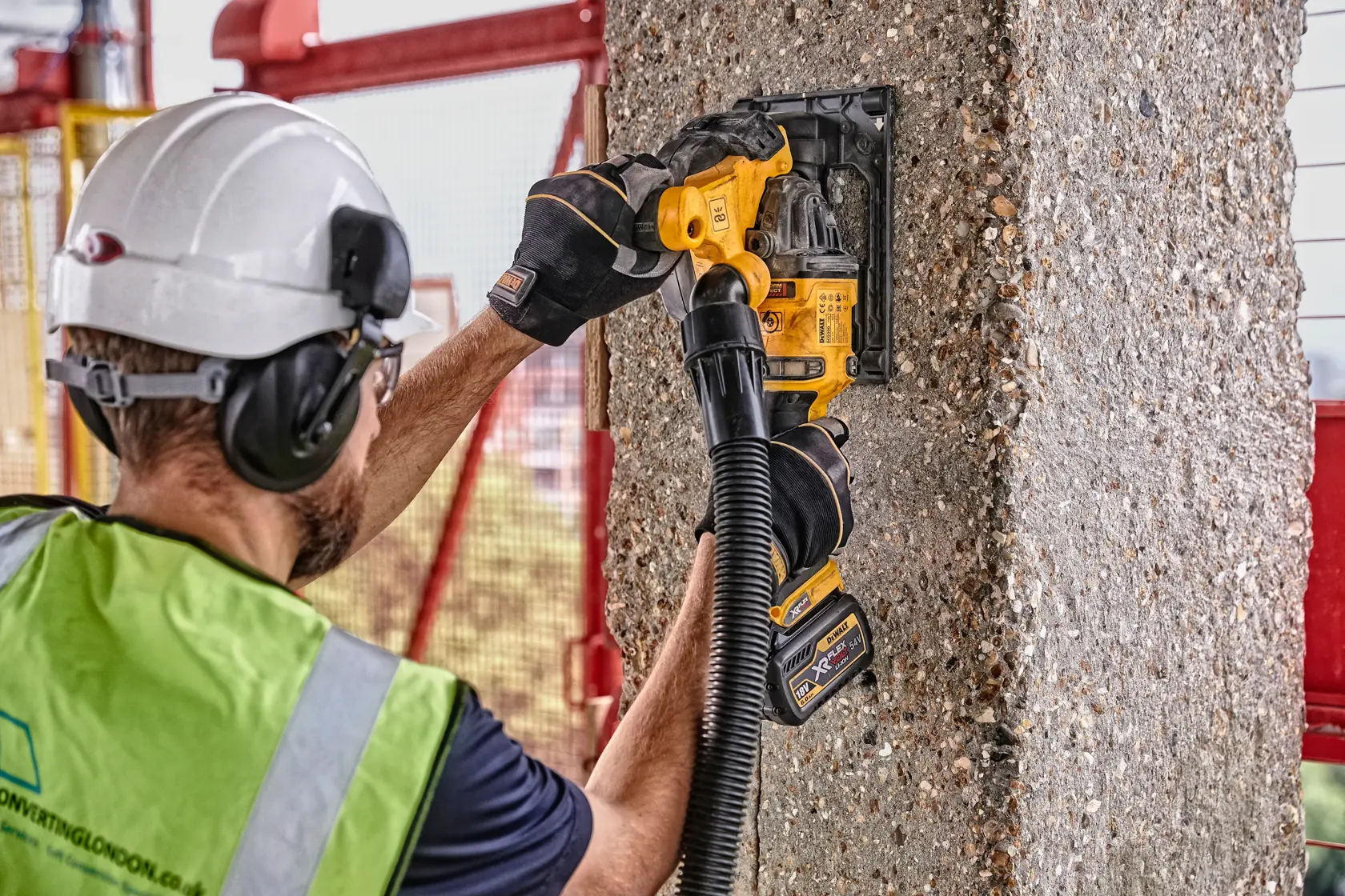 A worker wearing a safety helmet, gloves, and a high-visibility vest uses a DEWALT cordless tool to cut or grind into a concrete pillar. The tool appears to be fitted with a dust extraction hose for safety and cleanliness.