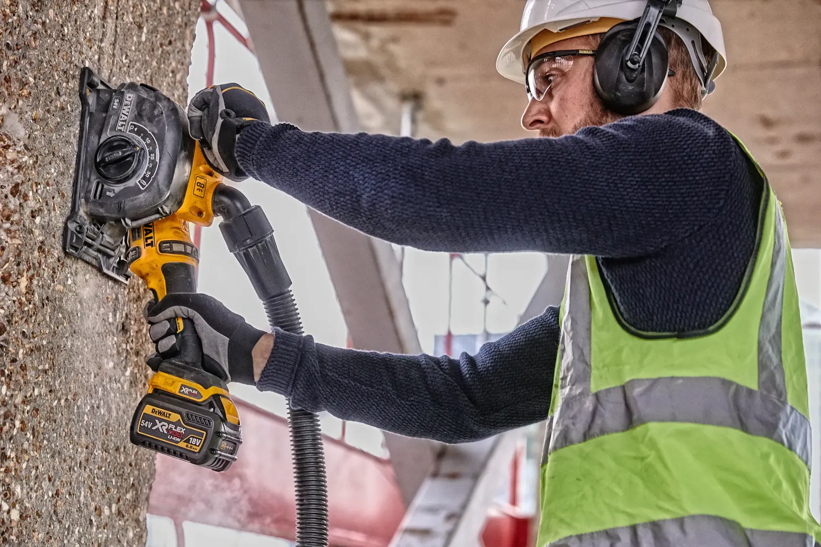 A person wearing safety gear uses a DEWALT cordless wall chaser tool to cut channels into a concrete wall at a construction site.