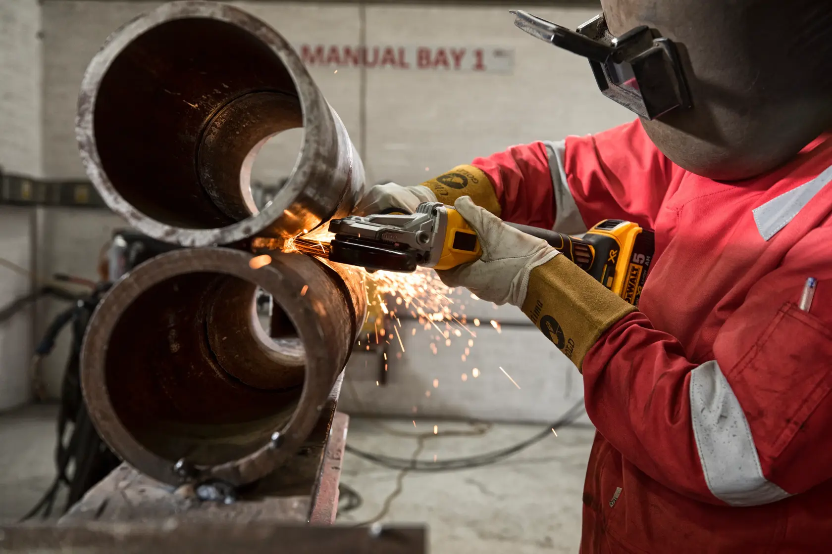 Worker using a DEWALT cordless grinder with sparks on metal pipes in an industrial workshop.