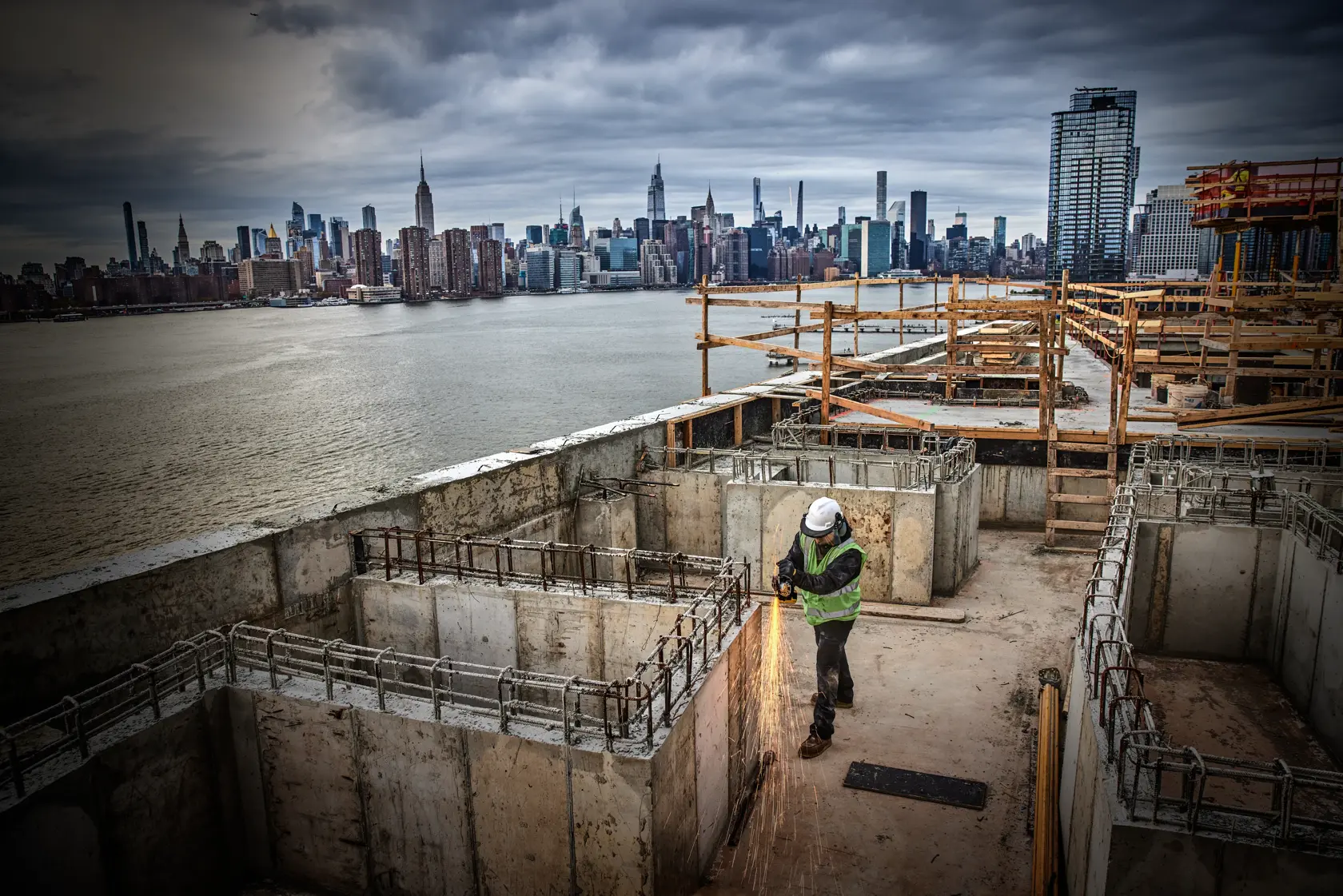 Worker uses power tool on concrete at construction site near waterfront, city skyline in background.