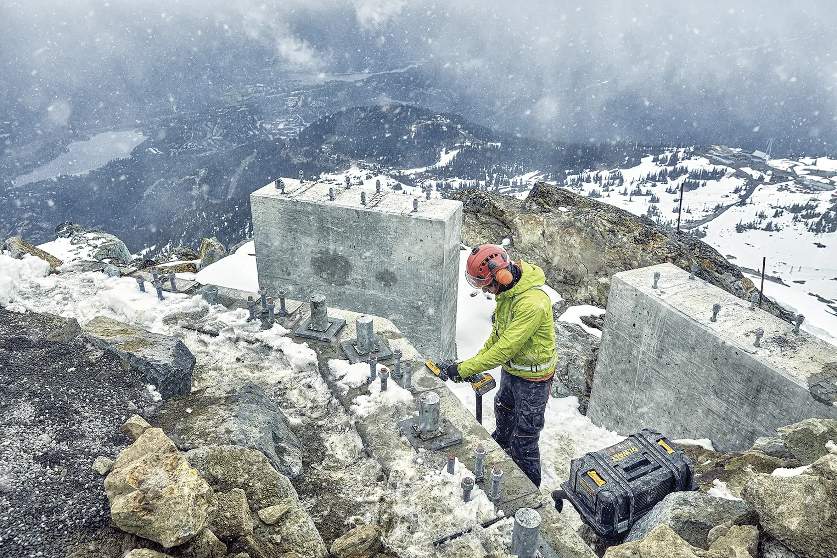Worker using DEWALT tool on snowy mountain construction site with concrete blocks and tool case.